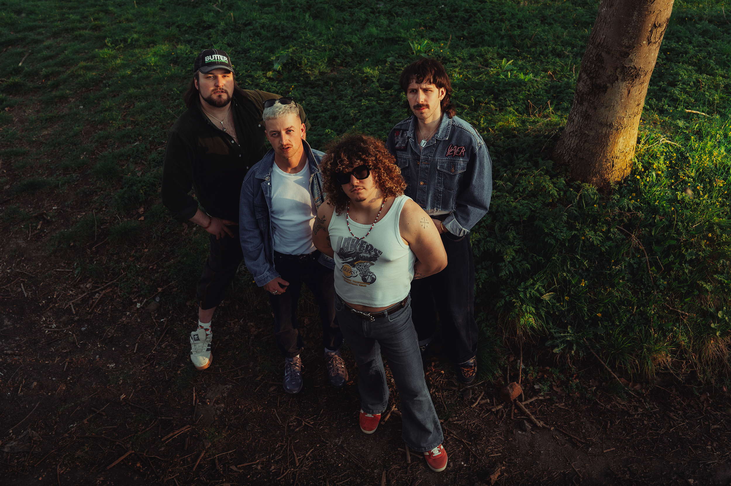 A group of five young men standing outdoors on grass and dirt, posing for a photo. They are looking up at the camera, with serious expressions. The setting appears to be natural with a large tree trunk on the right side.