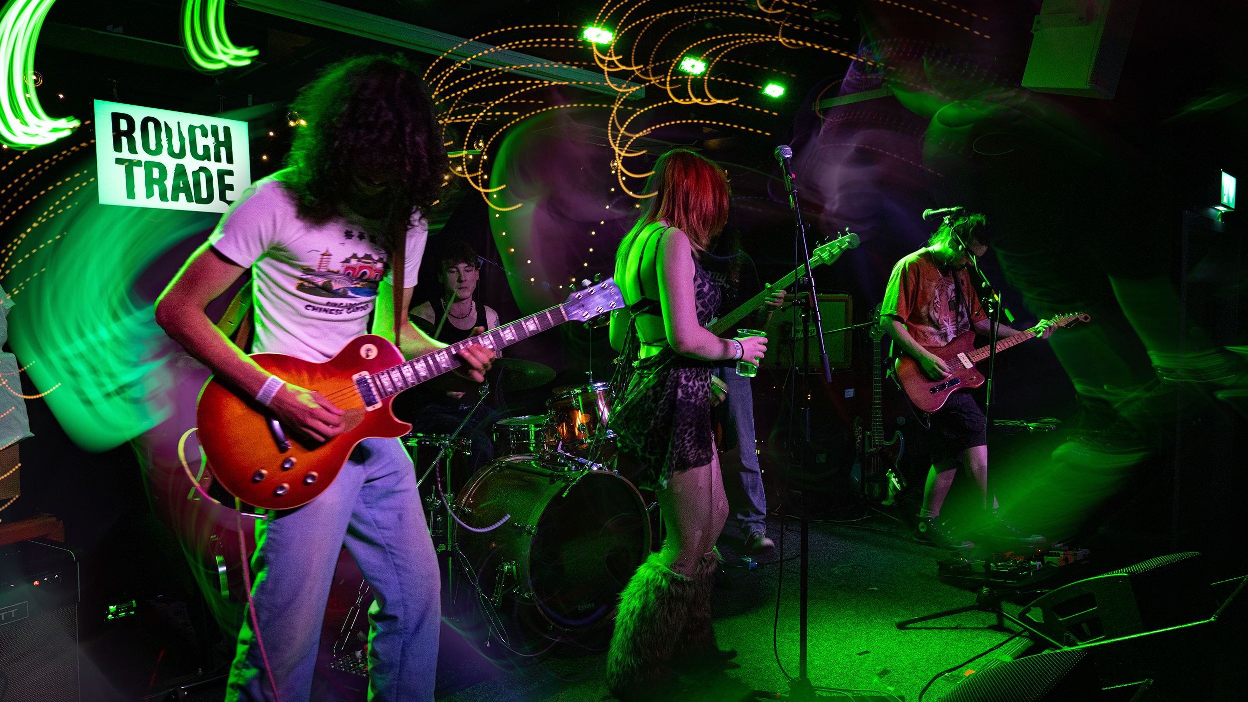 A band performs live on stage with colorful lighting, featuring multiple women playing guitars, a woman singing into a microphone, and a drummer in the background.