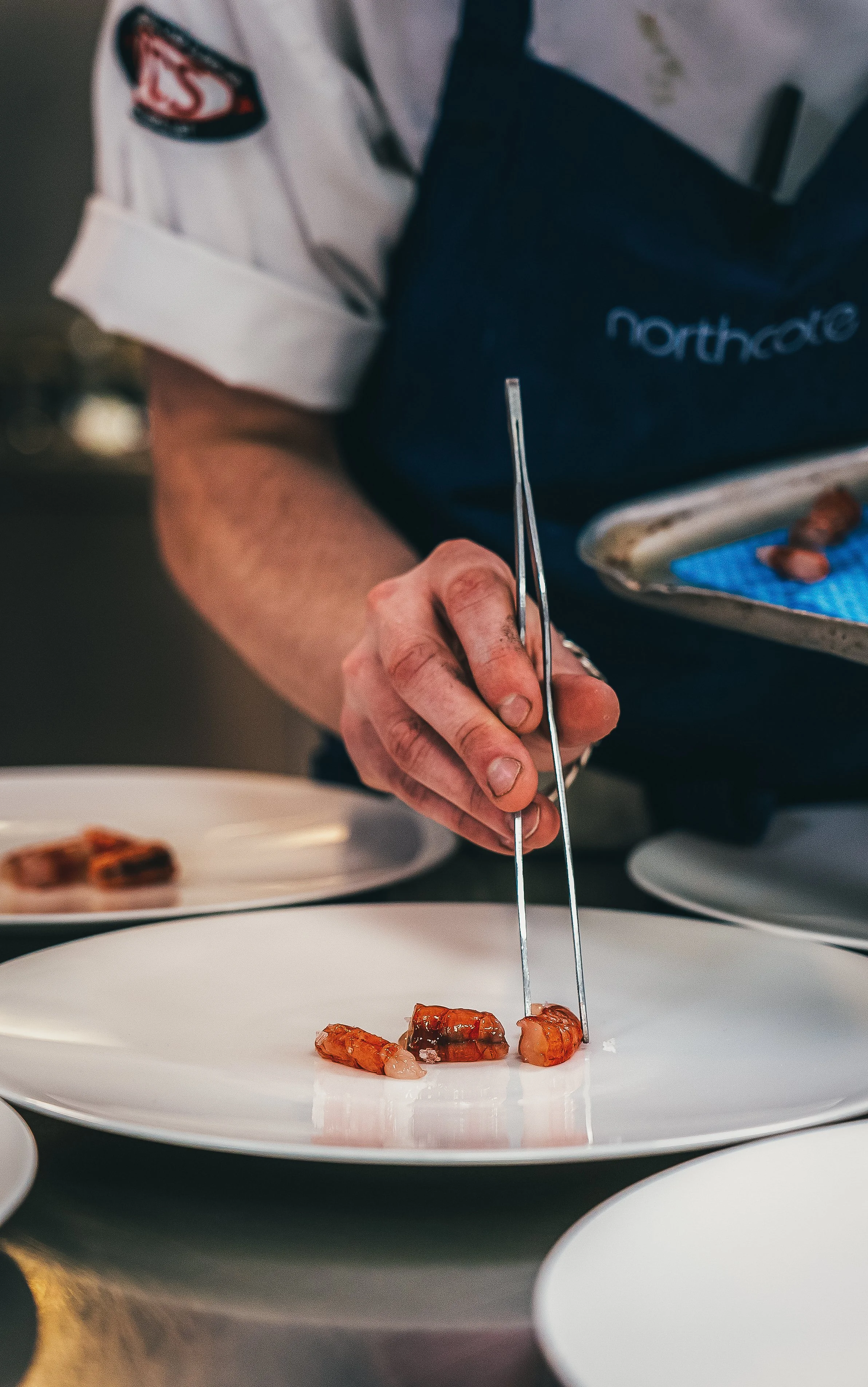 A chef using tweezers to place sashimi on a white plate, with a tray of sashimi behind him and plates on the table.