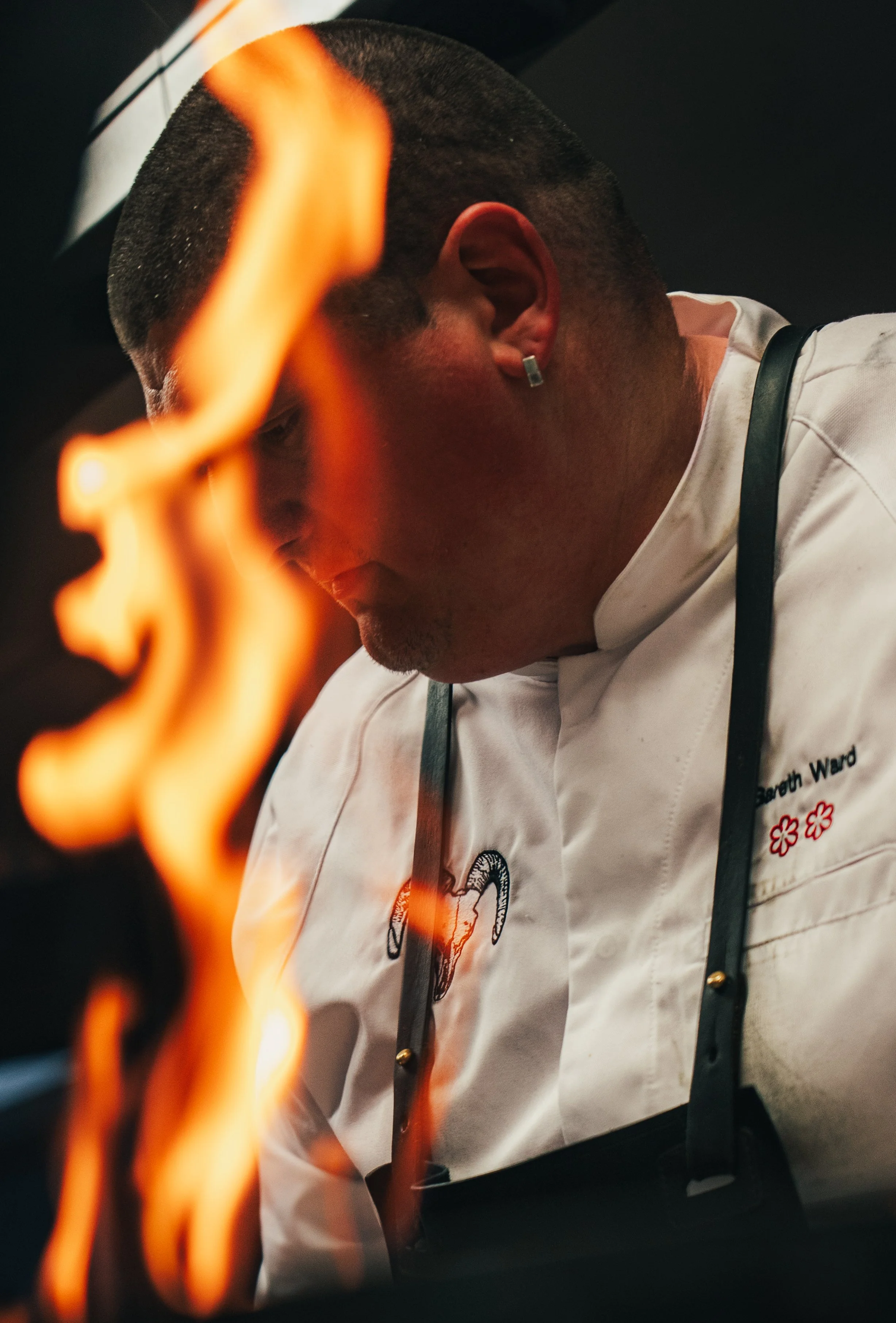 Close-up of a chef in a white uniform with embroidered red flowers, working near flames in a professional kitchen.