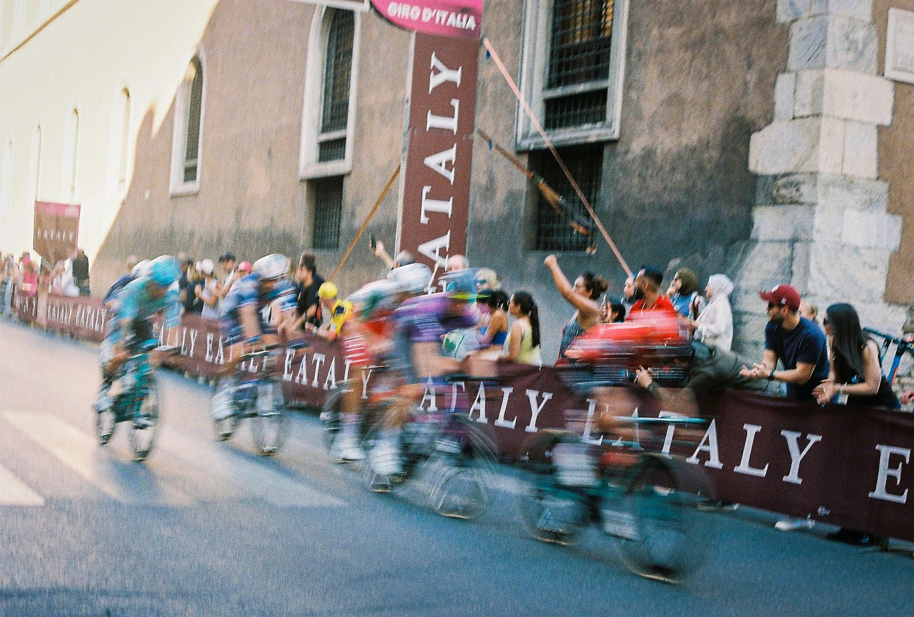 Bicycle race with multiple cyclists riding on a street in Italy, with spectators watching from the sidewalk and a sign with the word 'ITALY' on the fence.