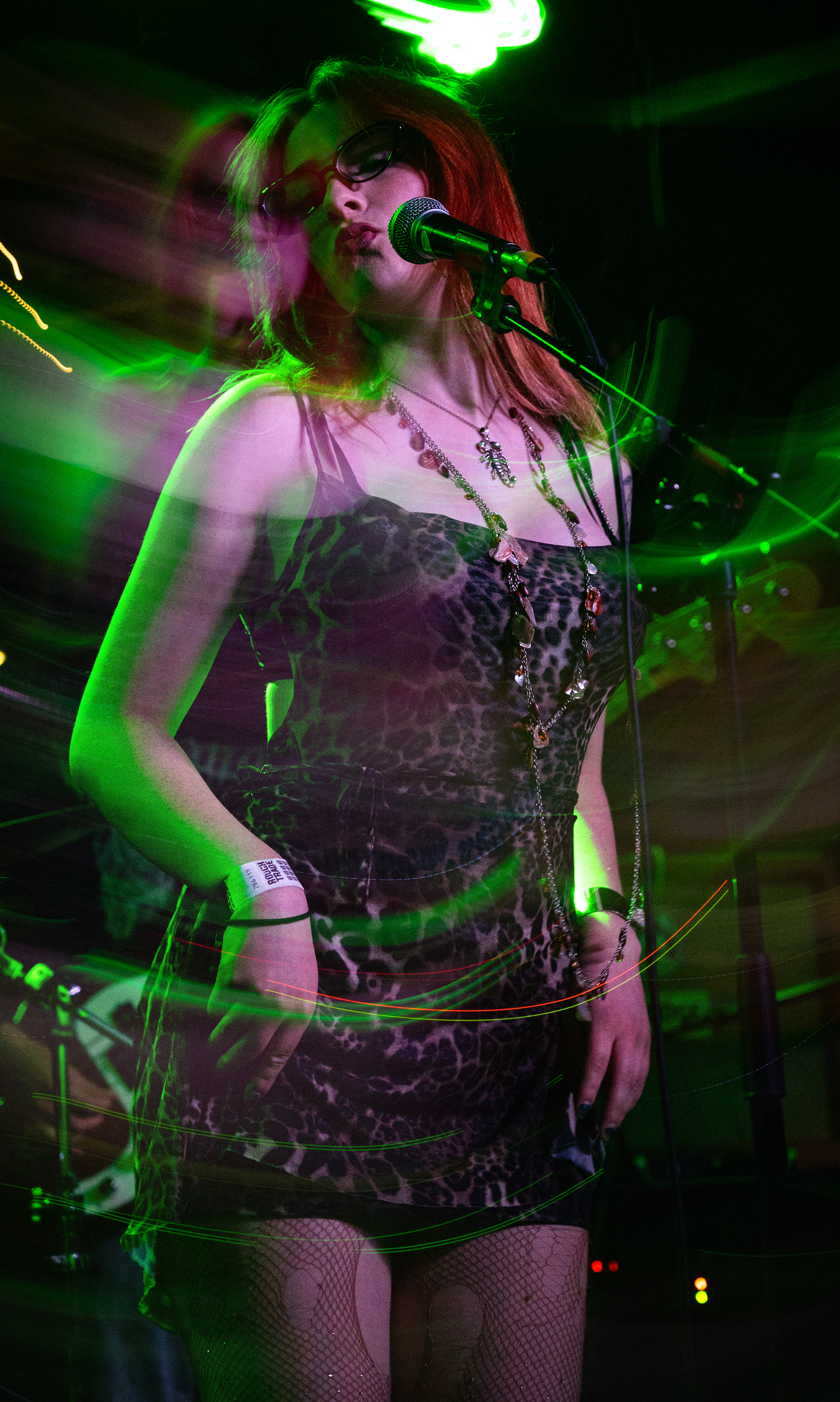 A woman with red hair, wearing sunglasses and a leopard print dress, singing into a microphone on stage surrounded by green lighting and motion effects.