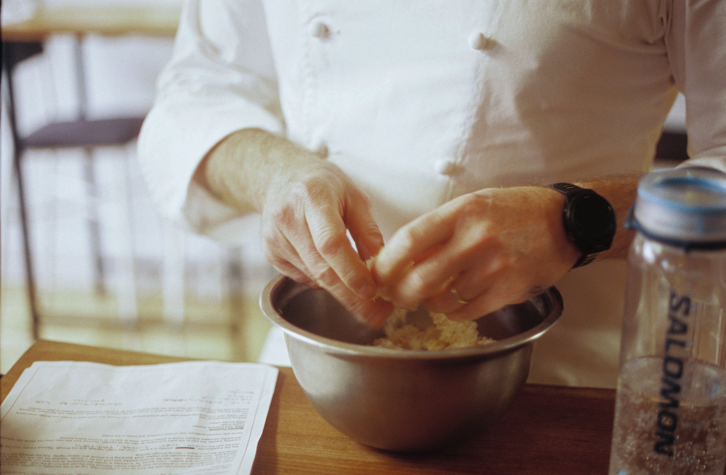 Person preparing batter with hands in a mixing bowl on a kitchen counter, surrounded by recipe paper and baking ingredients.