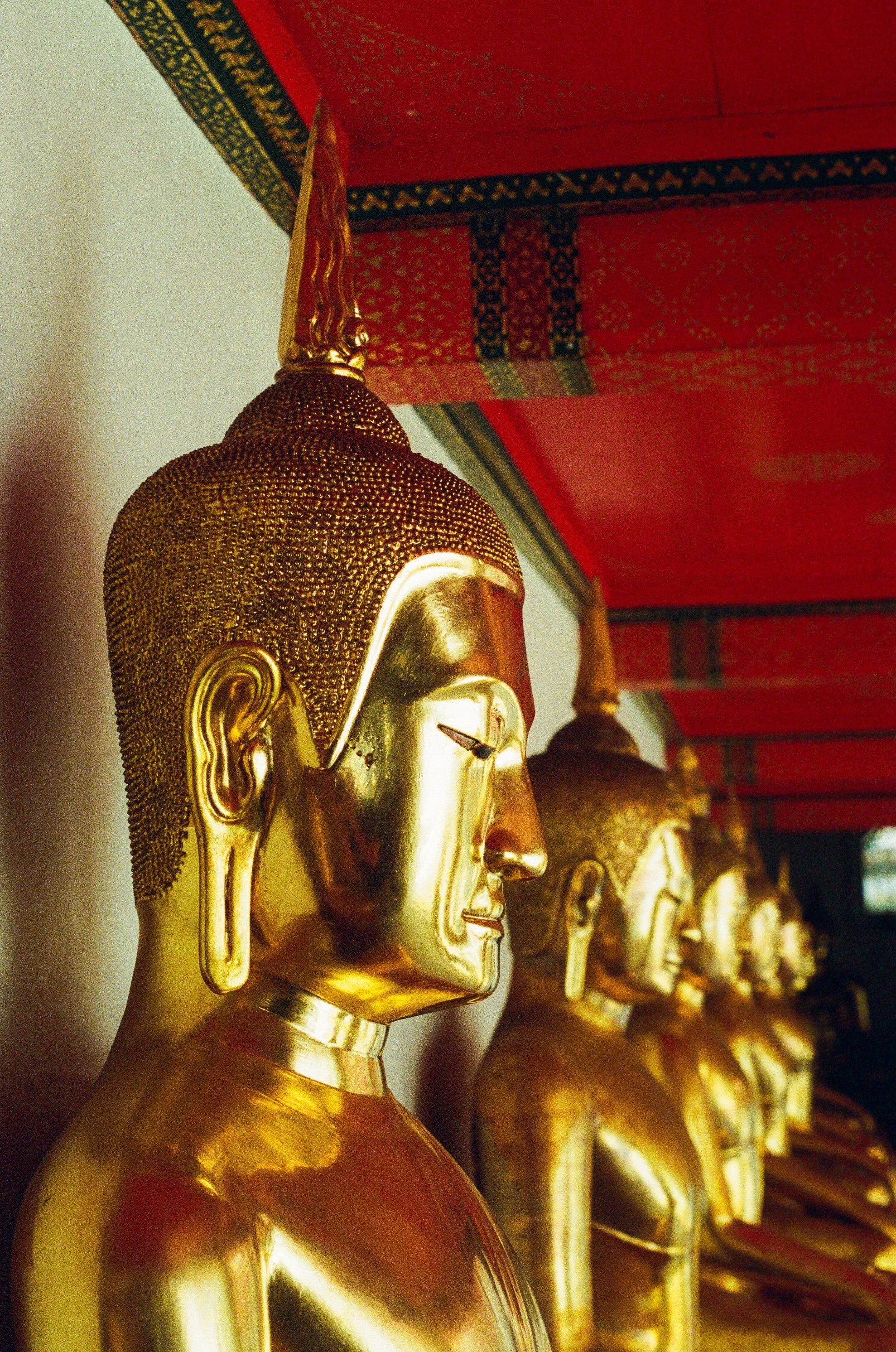 Multiple gold Buddha statues lined up under a red and gold decorated roof, viewed from the side.