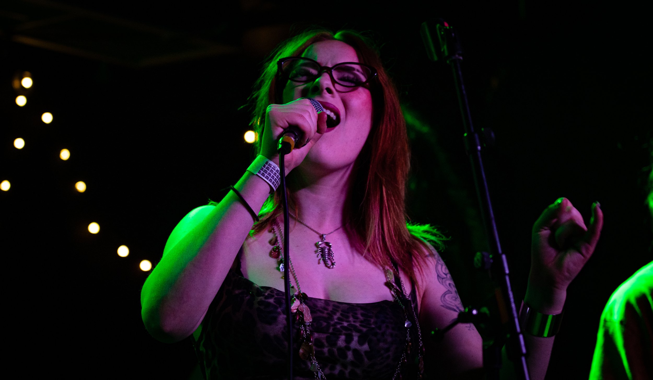 A woman with red hair and glasses singing into a microphone on stage, illuminated by green and purple lighting, with small yellow lights in the background.