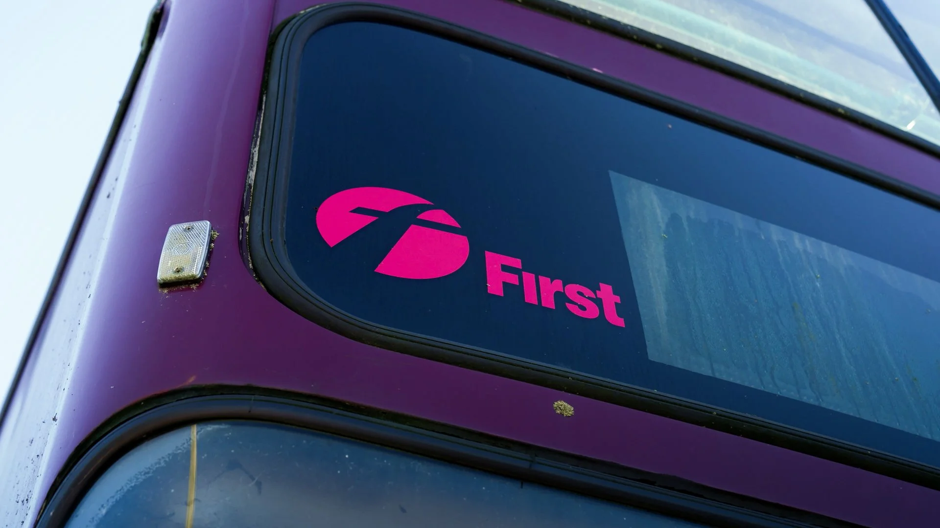A bus with a purple exterior displaying the FirstGroup logo and name on the window in pink, with part of the front and side of the bus visible.