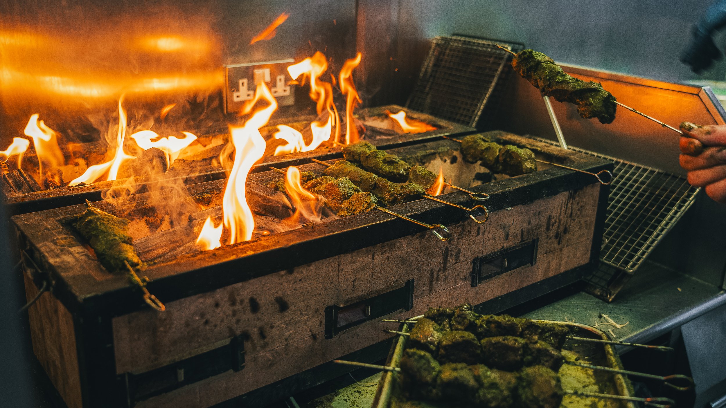 Skewers of marinated meat and vegetables being grilled over an open flame in a commercial kitchen.