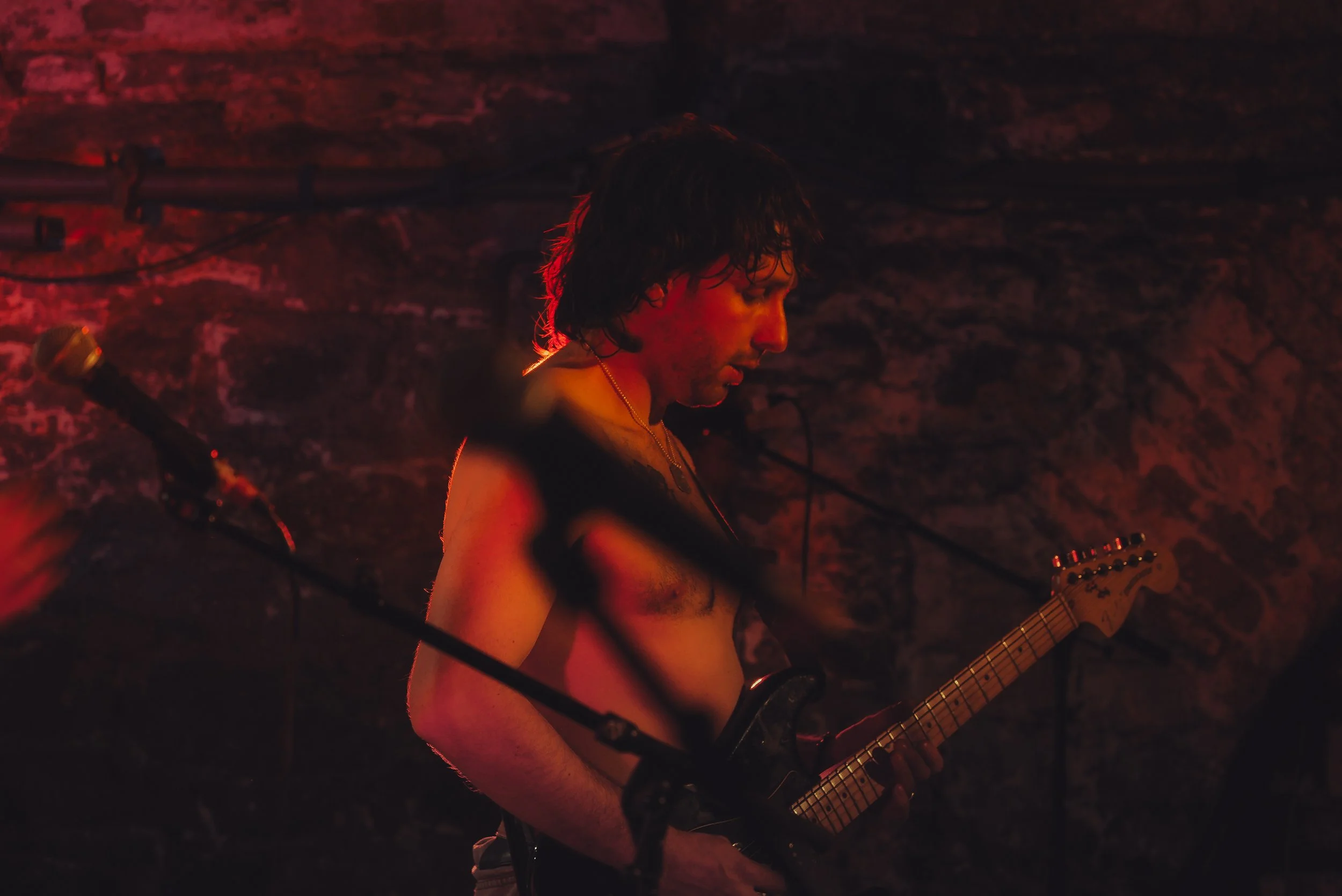 A man with long, dark, damp hair playing an electric guitar in a dim, red-lit setting with a exposed brick wall behind him.