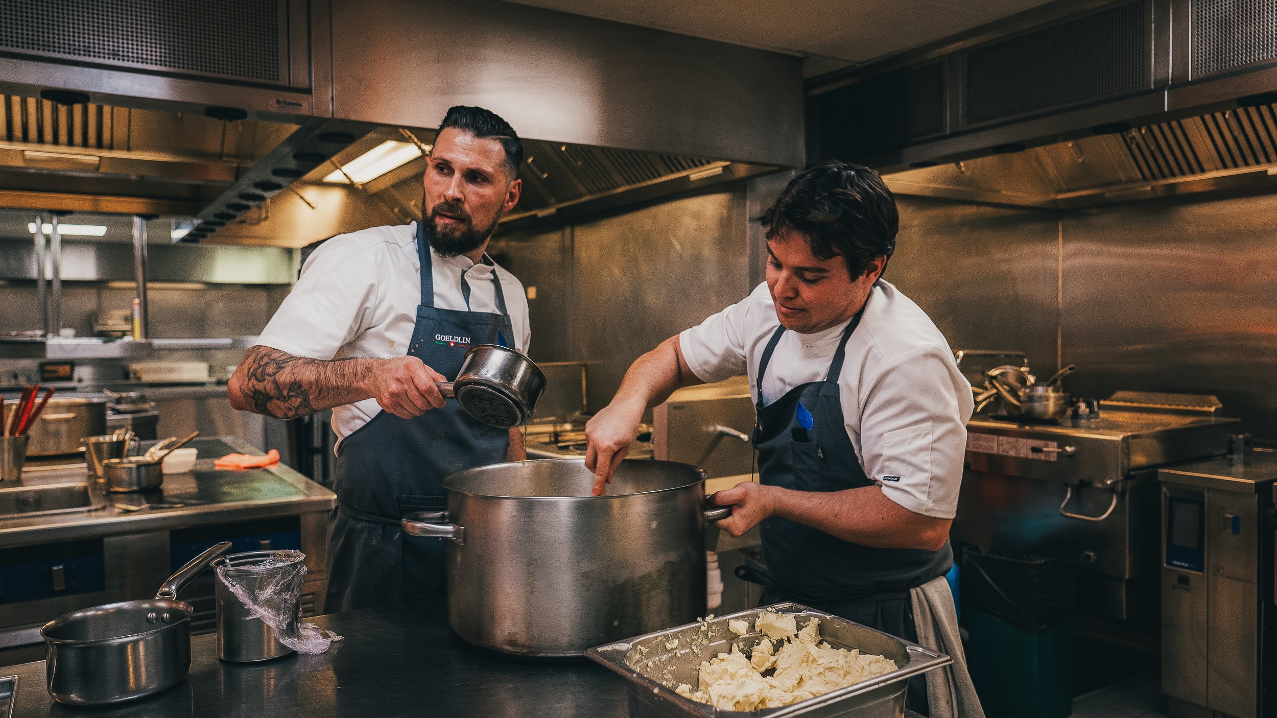 Two chefs working in a professional kitchen, preparing food together at a stainless steel counter.