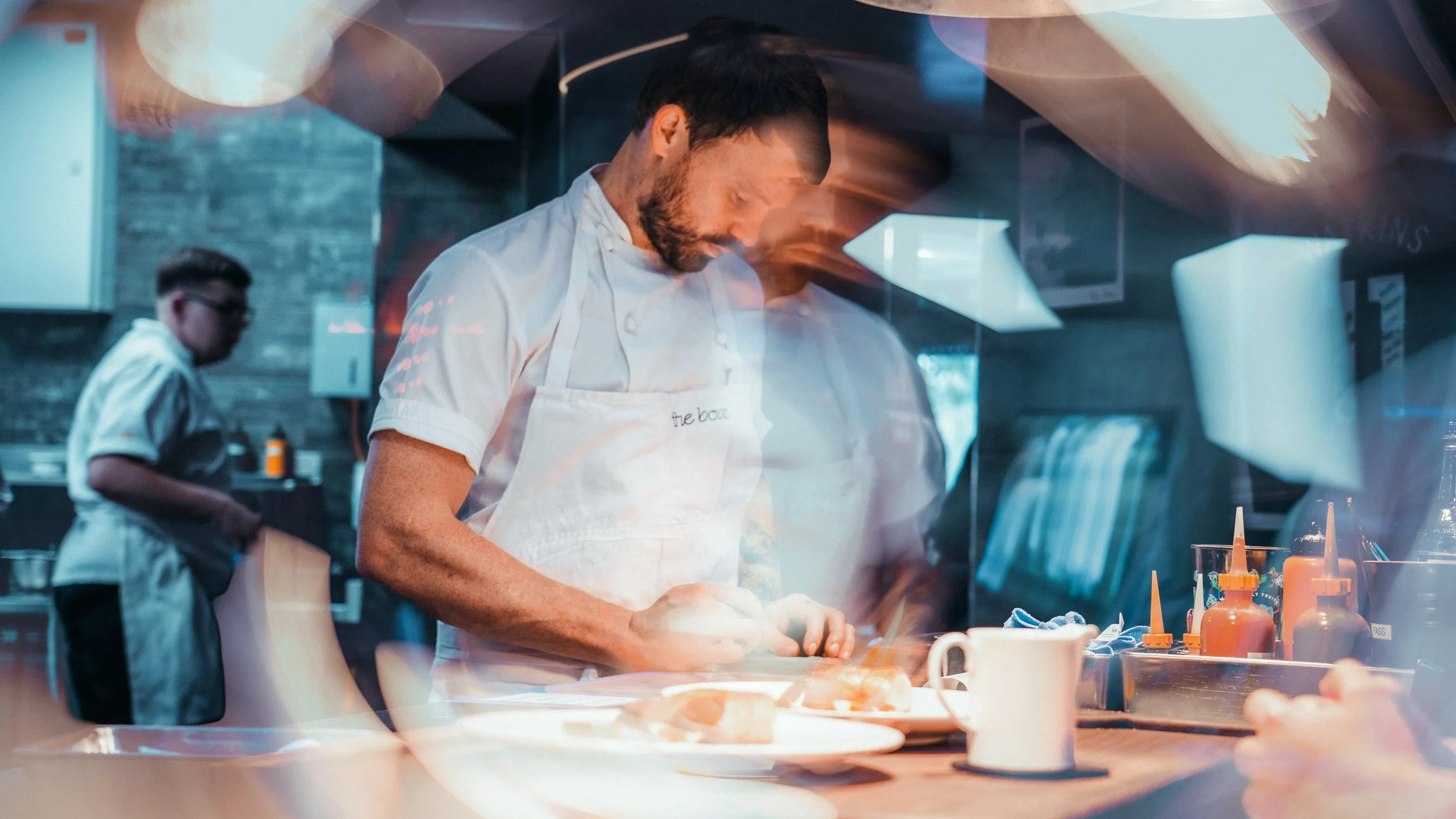 Chef in a white uniform preparing food in a modern kitchen with another chef in the background.