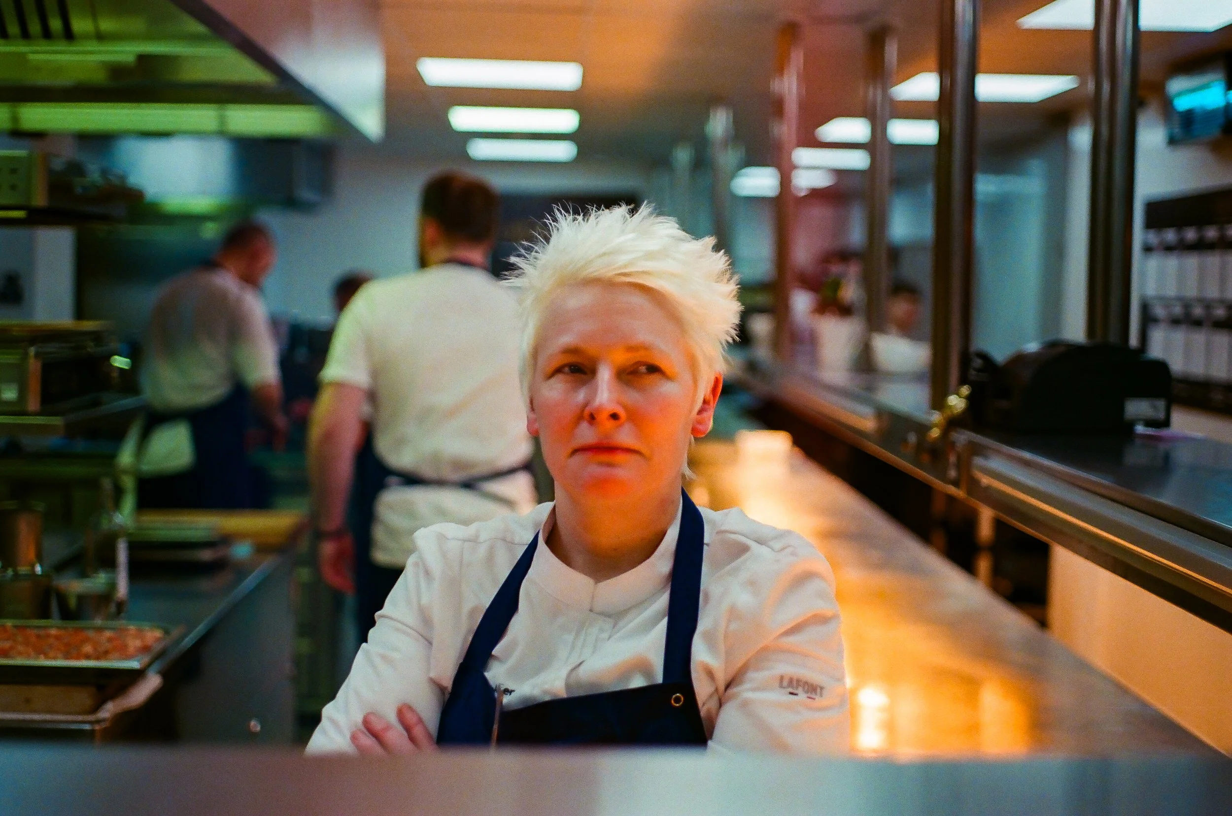Chef with platinum hair in a restaurant kitchen, standing with arms crossed, wearing a white chef's coat and blue apron, in front of a busy kitchen with other chefs working.