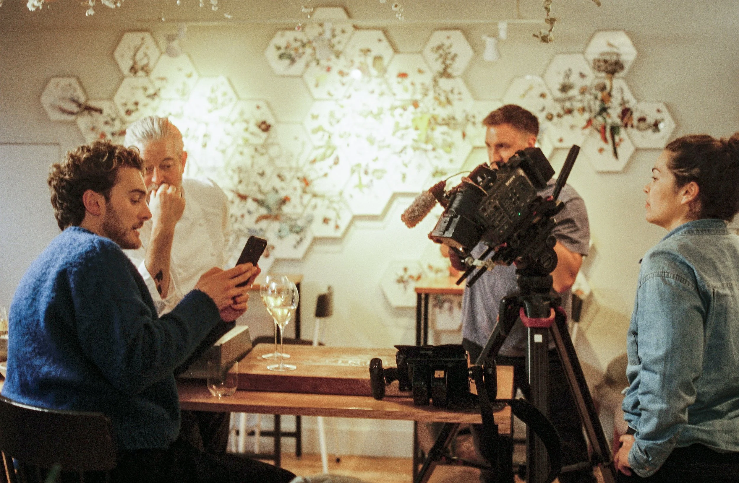 A group of four people in a restaurant or cafe setting, engaged in a conversation. One person is using a smartphone, a woman on the right is smiling, and a man is recording or preparing to record with a professional video camera. There is a decorative wall with honeycomb-shaped shelves behind them.