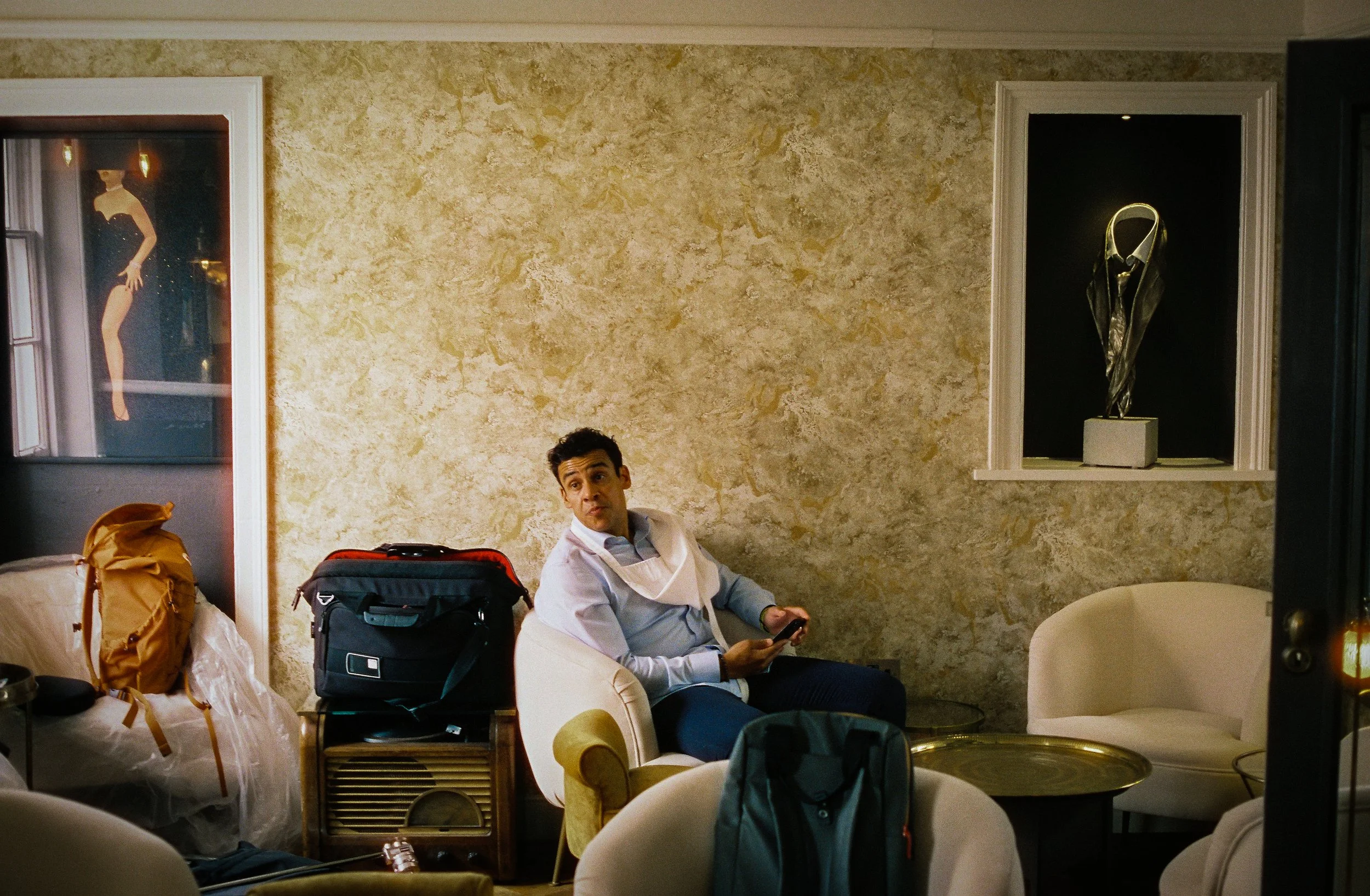 Man sitting on a beige armchair in a hotel room holding a phone, with luggage and packed bags around him, decorated with beige textured wall art and sculptures.