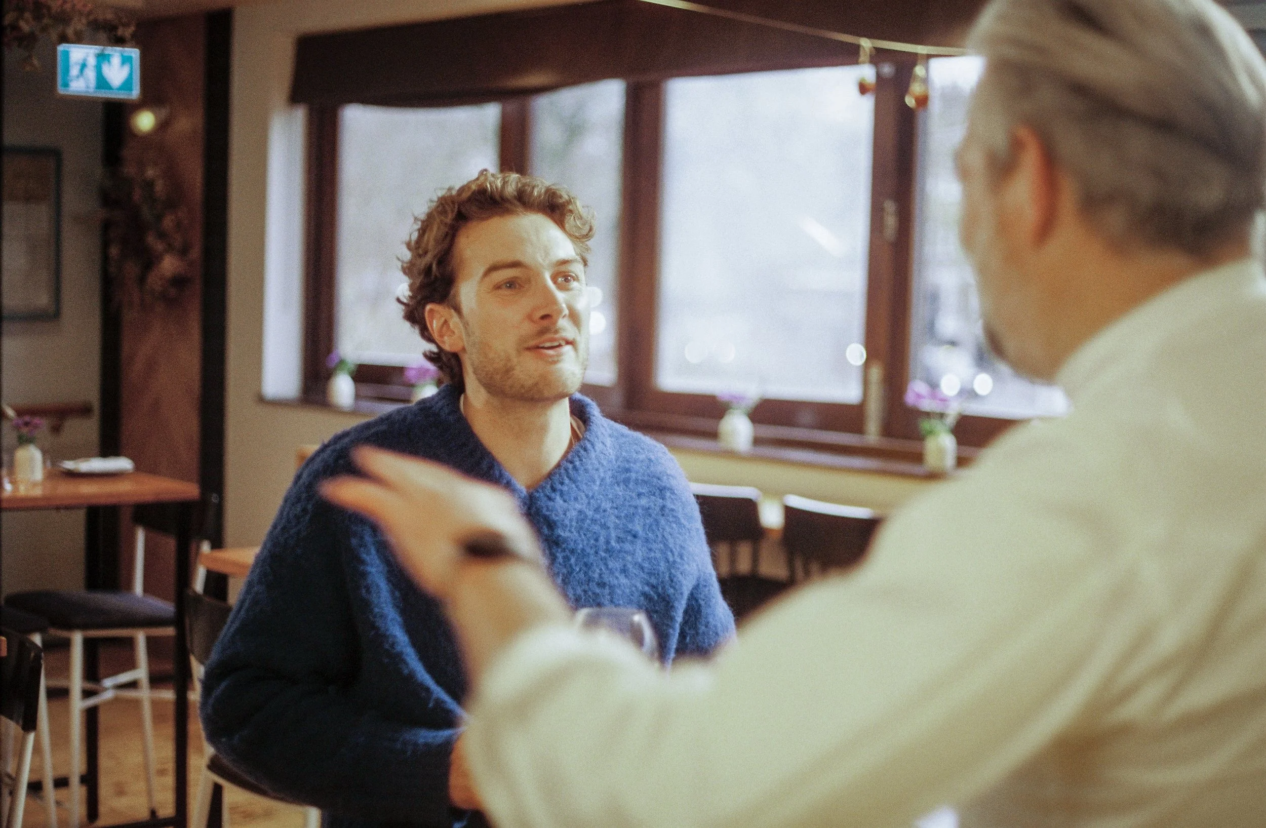 Two men engaging in a conversation inside a cozy cafe, one wearing a blue sweater and the other with gray hair. There are large windows with natural light, and a small vase with flowers on the windowsill.