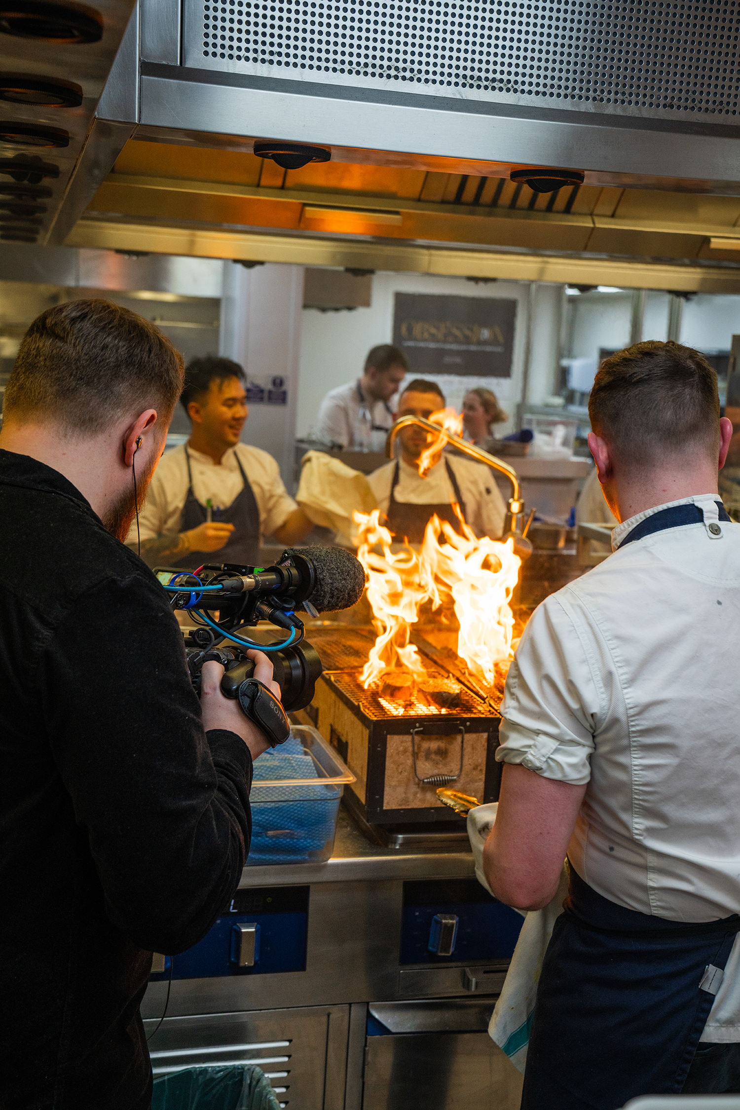 Chefs and videographer filming a chef cooking with flames in a commercial kitchen.