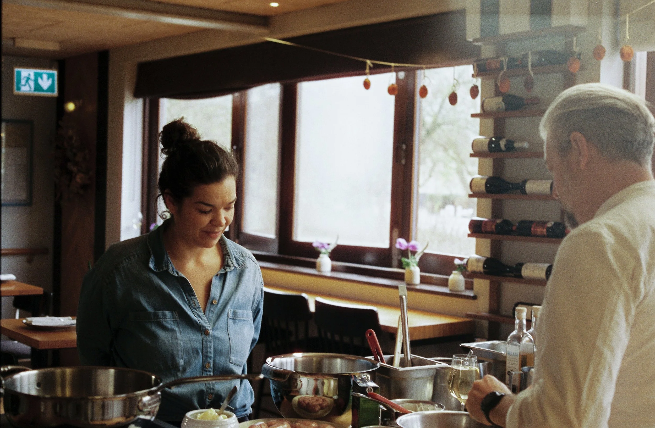Two people, a woman and a man, are preparing food in a cozy kitchen with wooden accents. The woman has dark hair tied up and is wearing a denim shirt, while the man has gray hair and is wearing a white shirt. They are working at a counter with various cooking ingredients and utensils, and wine bottles are displayed on shelves in the background. Sunlight is coming through the window, creating a warm atmosphere.