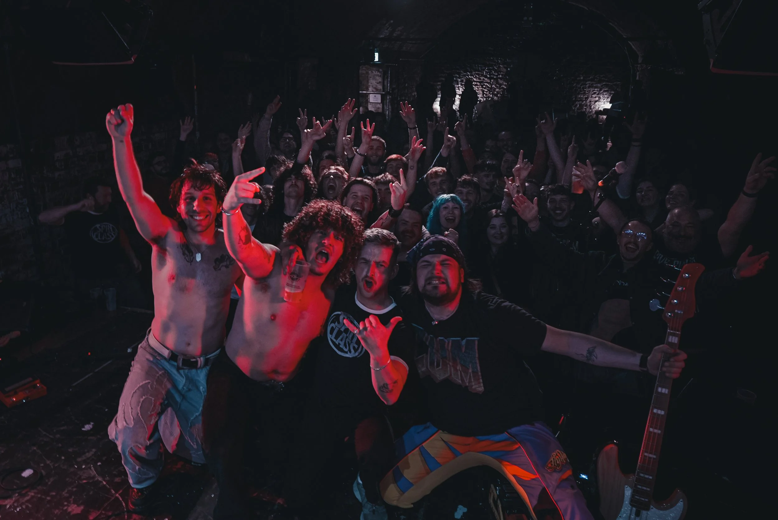 Group of people at a concert or music venue, some smiling and making gestures, with band equipment and a dark brick wall in the background.