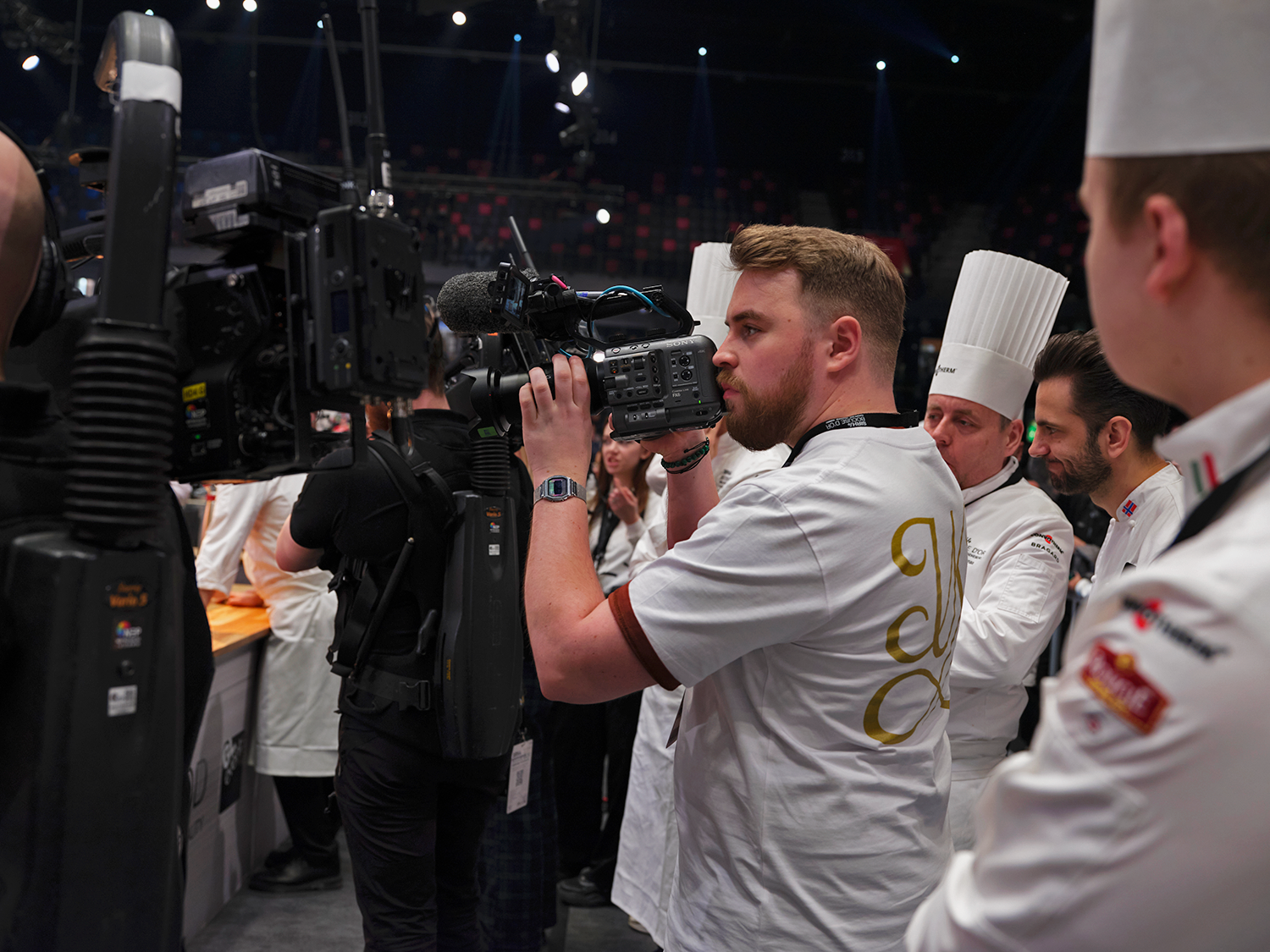 A man is filming with a professional video camera during a culinary event, with chefs and other participants in the background.
