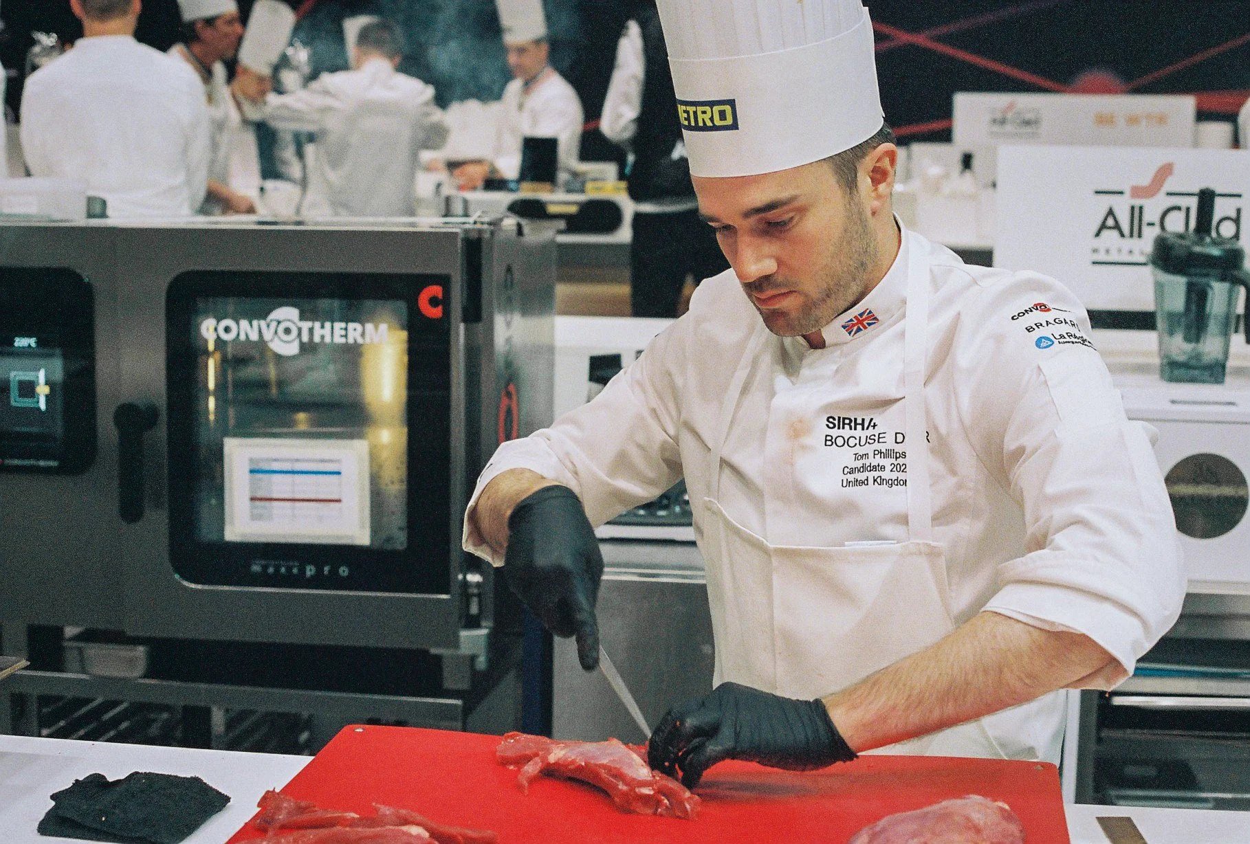 A chef wearing a white uniform and tall chef hat, with a British flag pin, is preparing meat on a red cutting board in a professional kitchen. Several other chefs are working in the background, and there is a black convection oven with the brand name 'CONVOTHERM' in front of him.