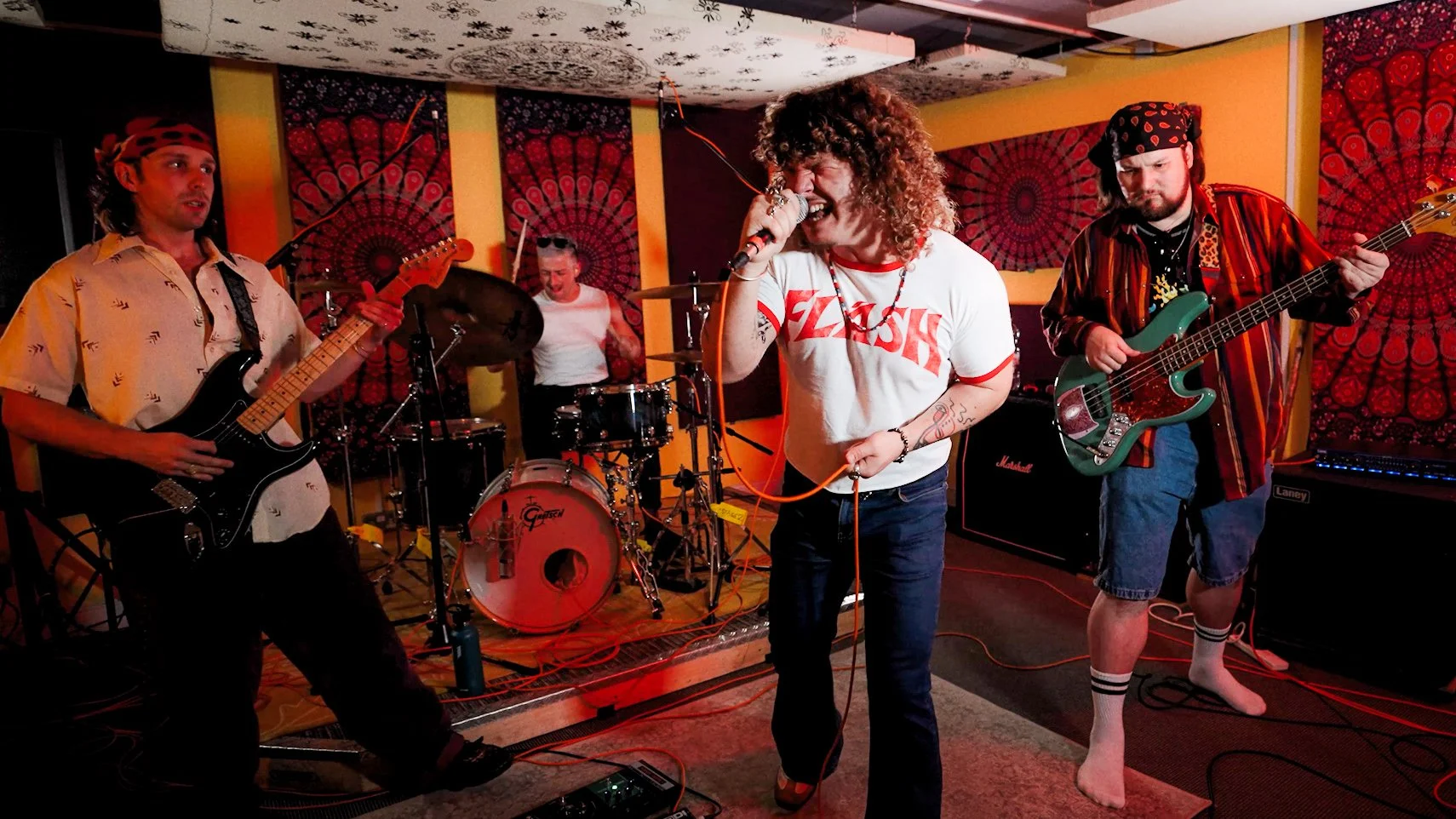 A band of four musicians performing in a vibrant, colorful venue. The lead singer has curly hair and is passionately singing into a microphone, wearing a white T-shirt that says 'FLEX' and dark jeans. To the left, a guitarist in a patterned shirt and headband plays an electric guitar. Behind the singer, a drummer plays drums, and to the right, a bassist in a red and black striped shirt and shorts plays an electric bass guitar. The background features red and yellow patterned curtains, creating a lively atmosphere.