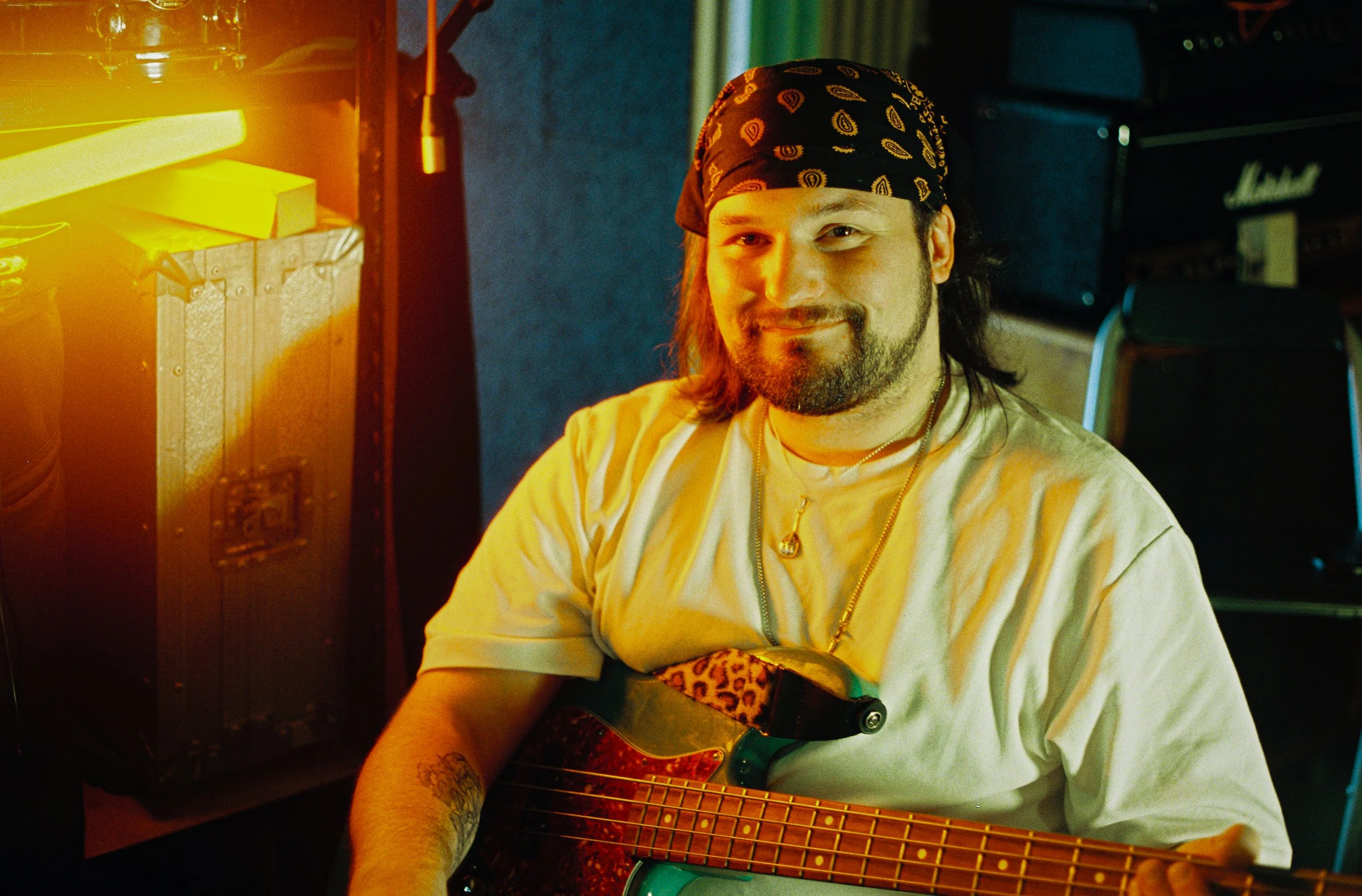 A man with a bandana on his head, smiling while holding an electric guitar, sitting in a dimly lit room with musical equipment around him.