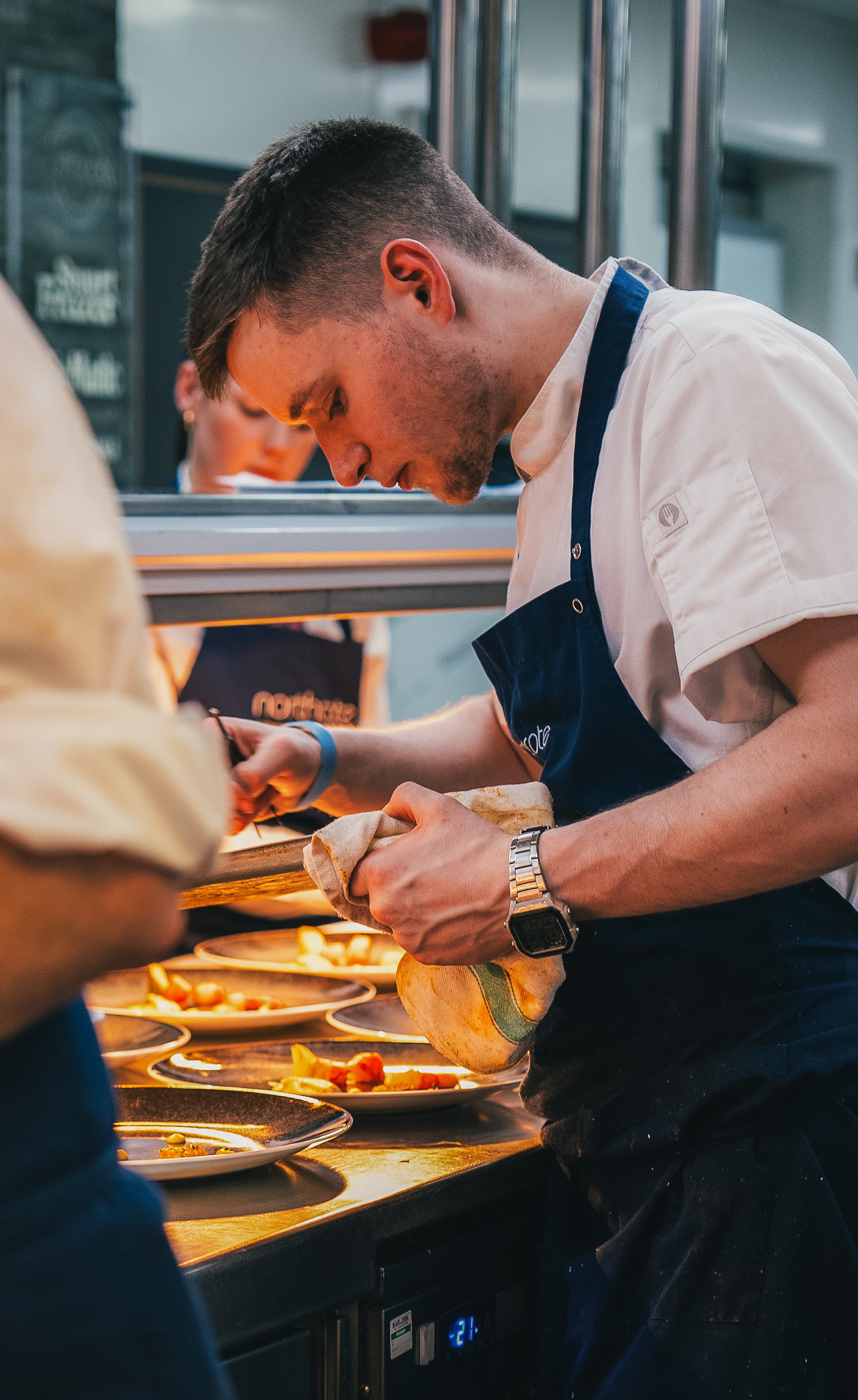A young male chef wearing a white shirt and a dark apron is preparing food in a professional kitchen. He is focused on his task, holding a cloth to handle a kitchen towel or dish. Plates of prepared food can be seen on the counter in front of him, an
