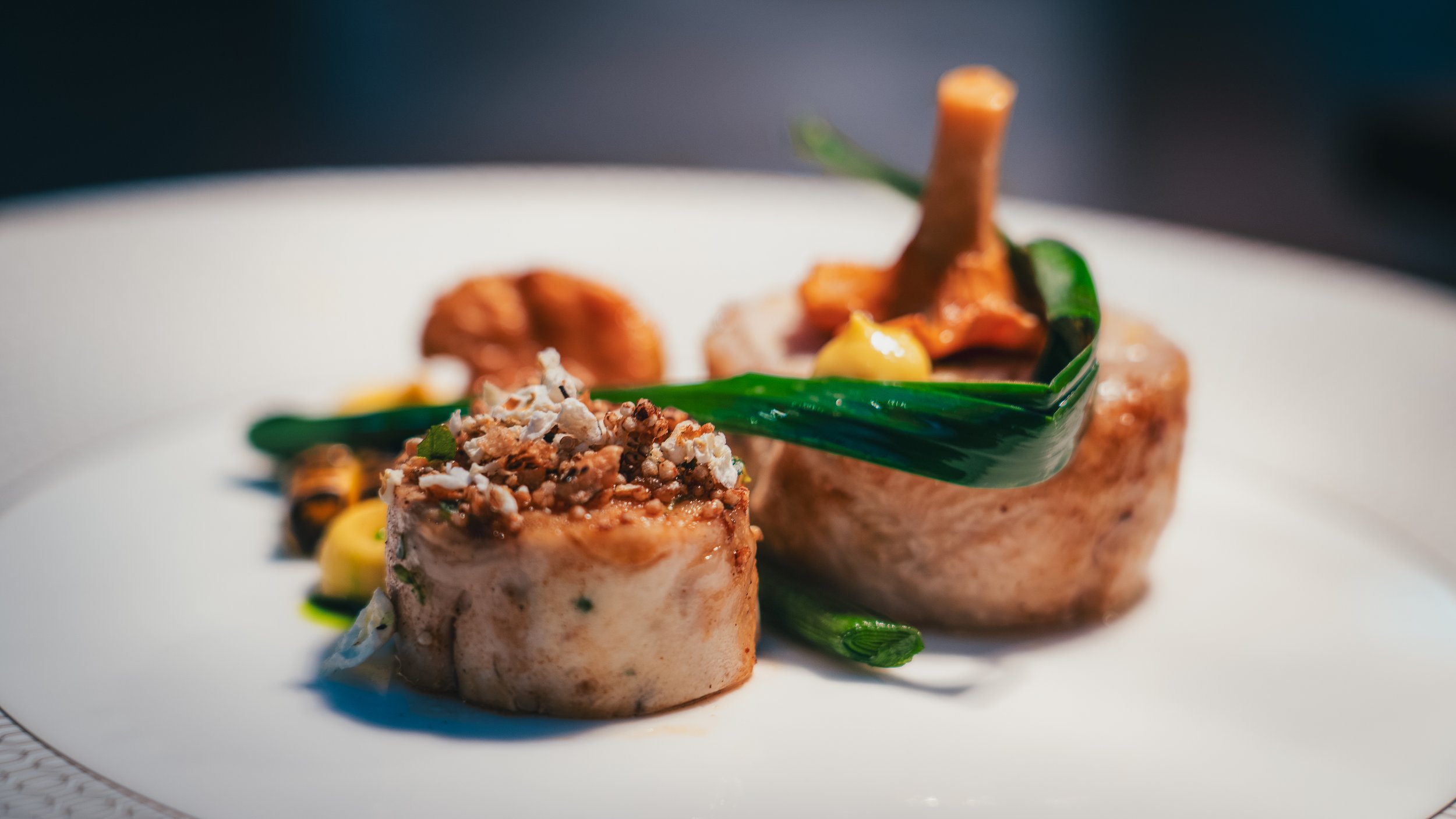 Two intricately plated gourmet dishes featuring seared seafood topped with crunchy garnishes, accompanied by green vegetable garnishes on a white plate.
