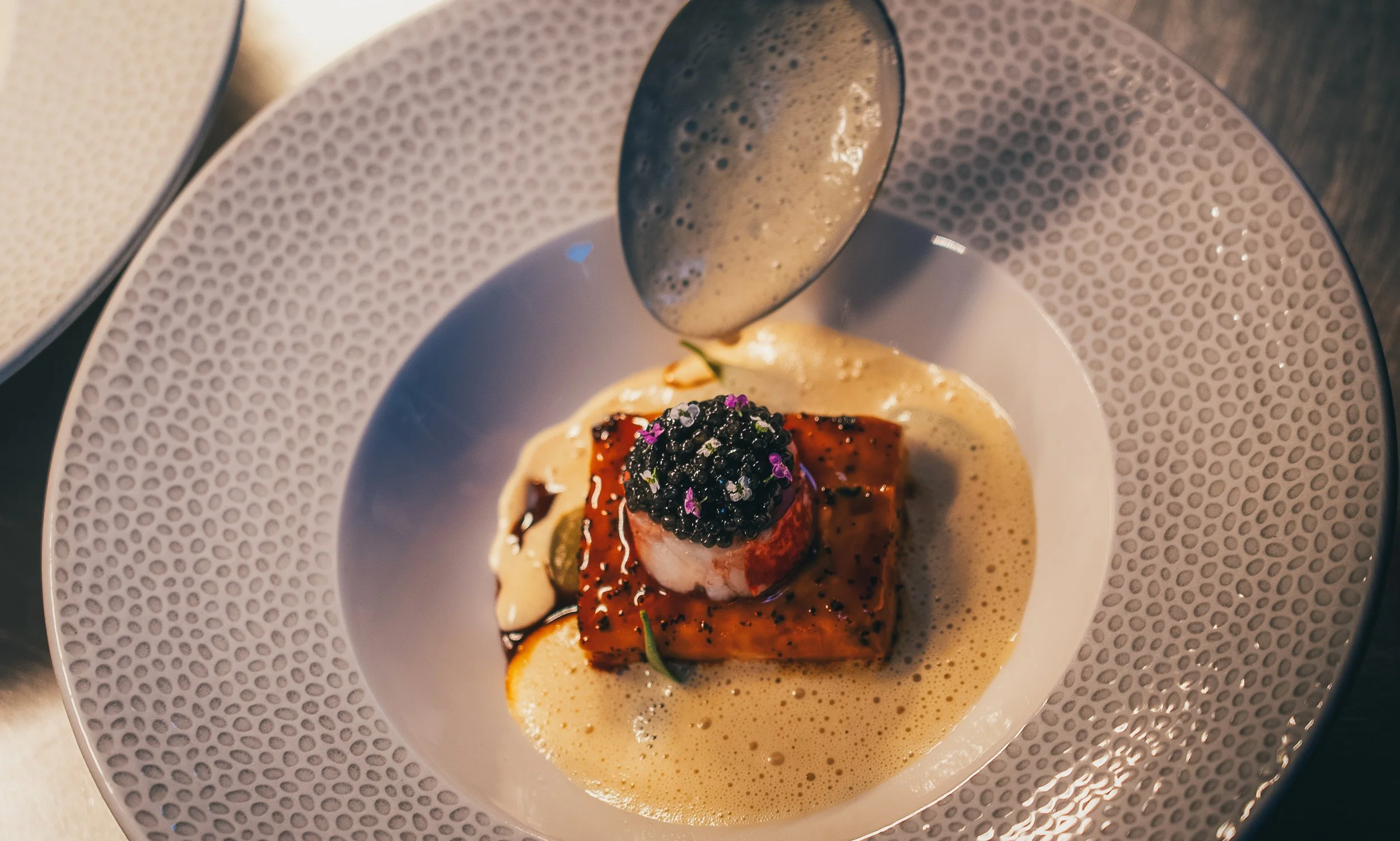 A plated gourmet dish featuring a seared piece of meat topped with black caviar and purple edible flowers, served with a creamy sauce and garnished with microgreens, on a decorative patterned plate.