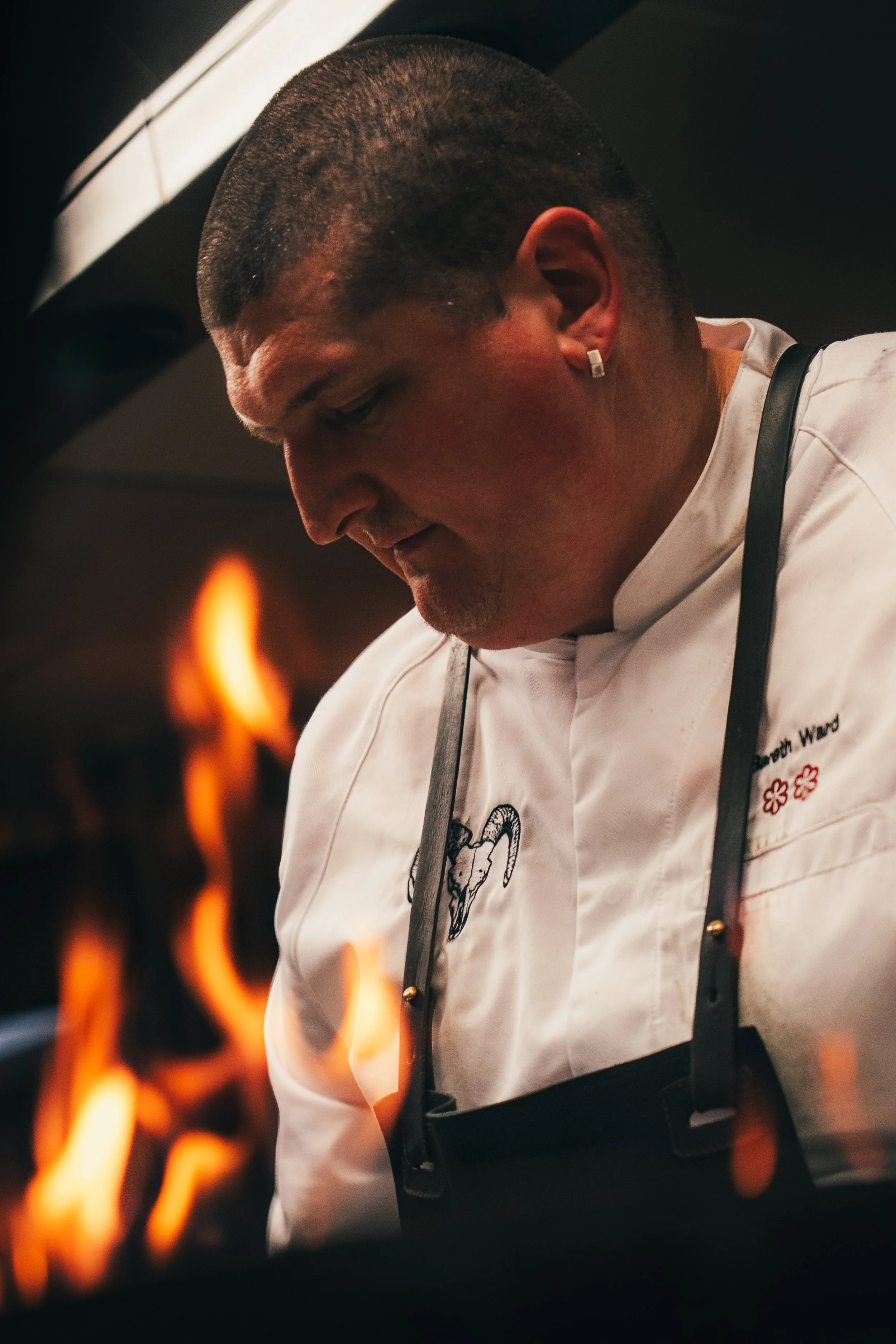 Close-up of a chef in a white uniform with a goat illustration on the chest, wearing a black apron and earrings, working near open flames in a kitchen.