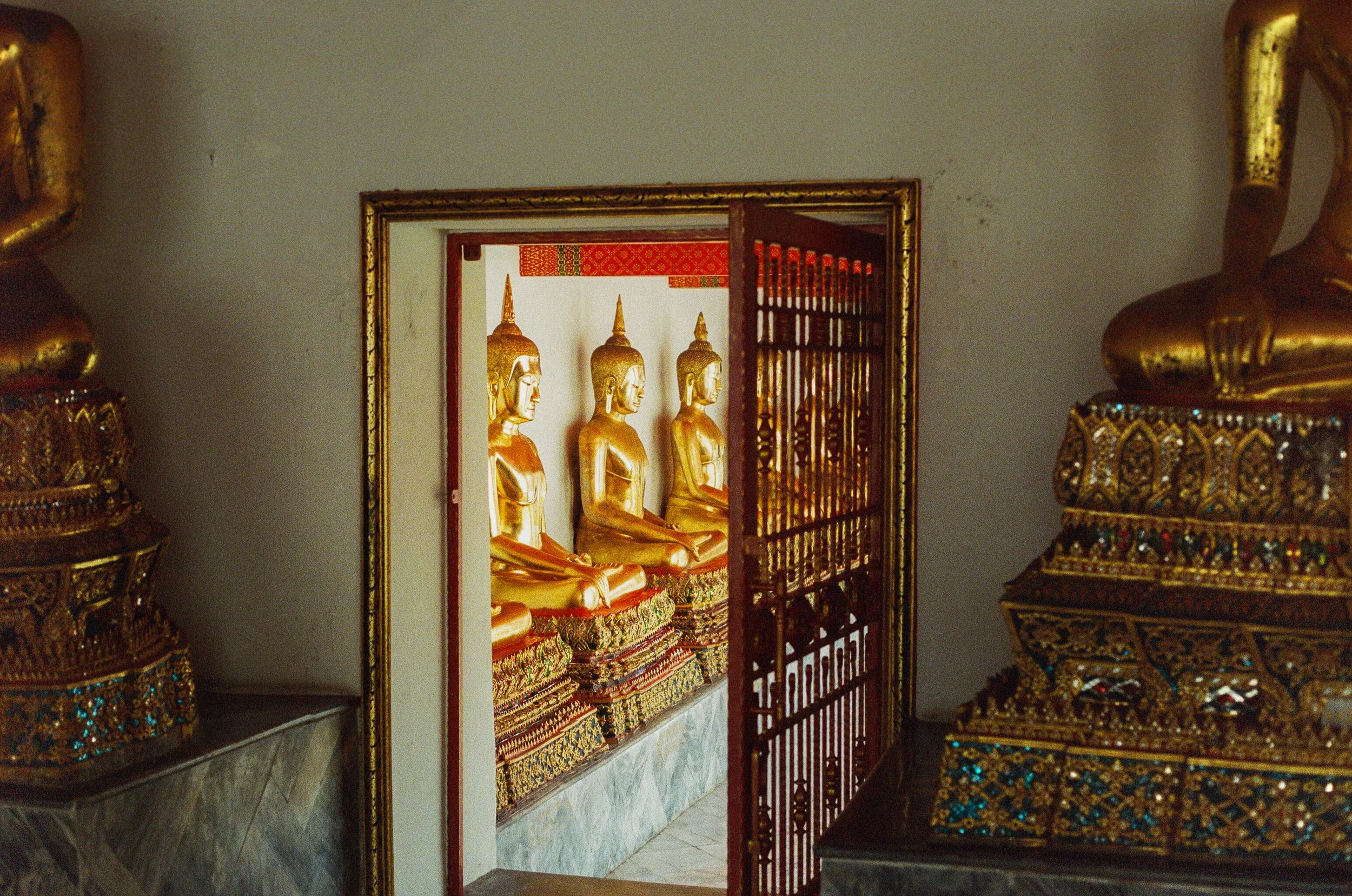 Golden seated Buddha statues in a temple room, framed by decorative structures and ornate details, with a red and gold patterned roof visible in the background.