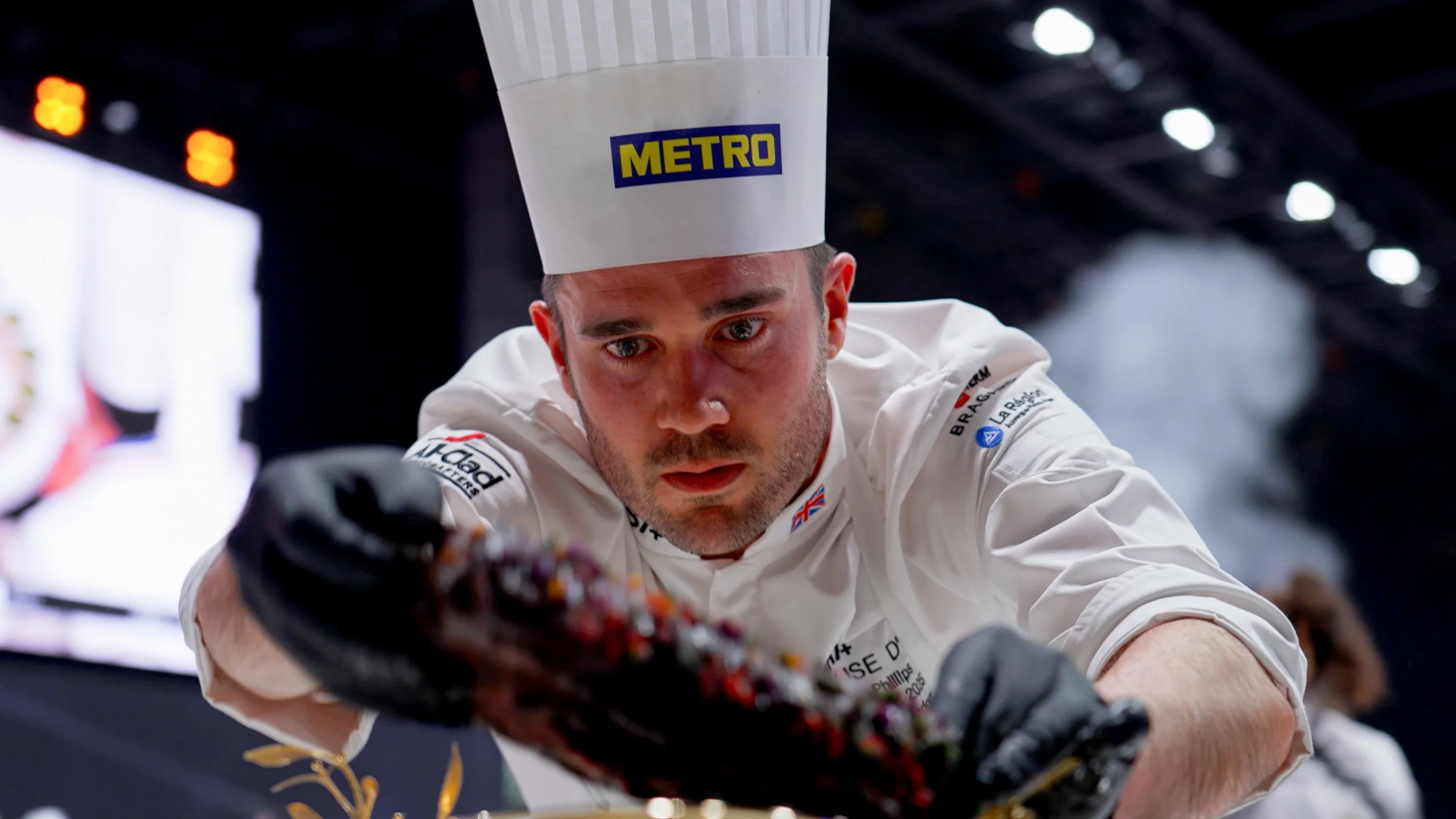 A chef in a white uniform and chef hat with a 'METRO' logo inspects a dish, focusing intently on it.