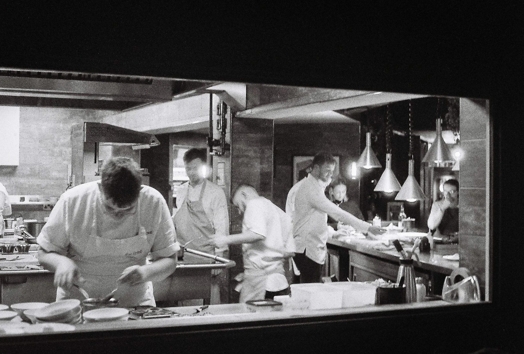 Black and white photo of a busy restaurant kitchen with chefs preparing food, seen through a window.