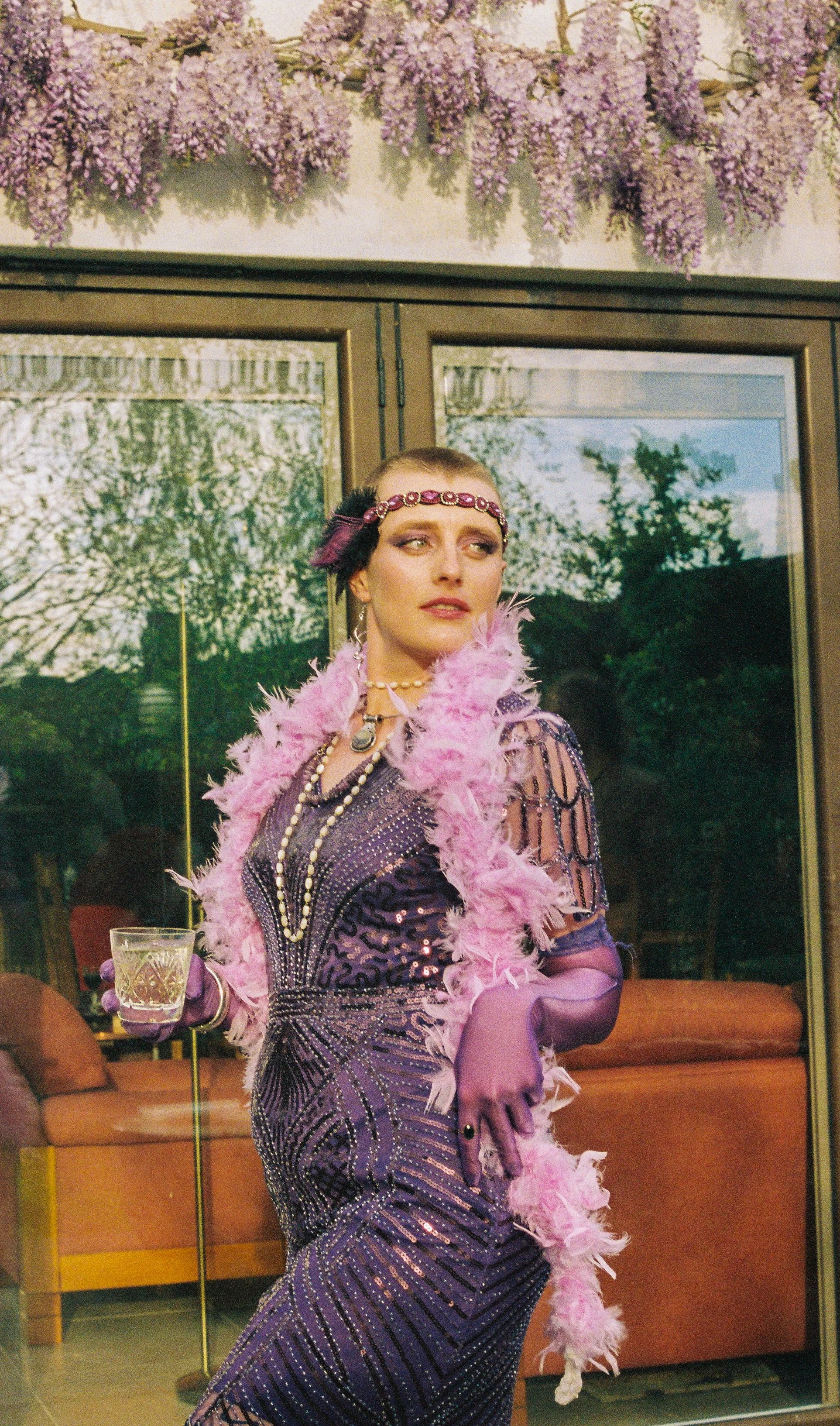 A woman dressed in an elegant purple, glittering, beaded gown with many layers of jewelry, including pearl necklaces and a gem bracelet, wearing a pink feather boa and purple gloves, holds a glass of drink. Her hair is adorned with a bead headband and feathers. She stands indoors near glass doors with trees visible outside and purple flowers hanging above.