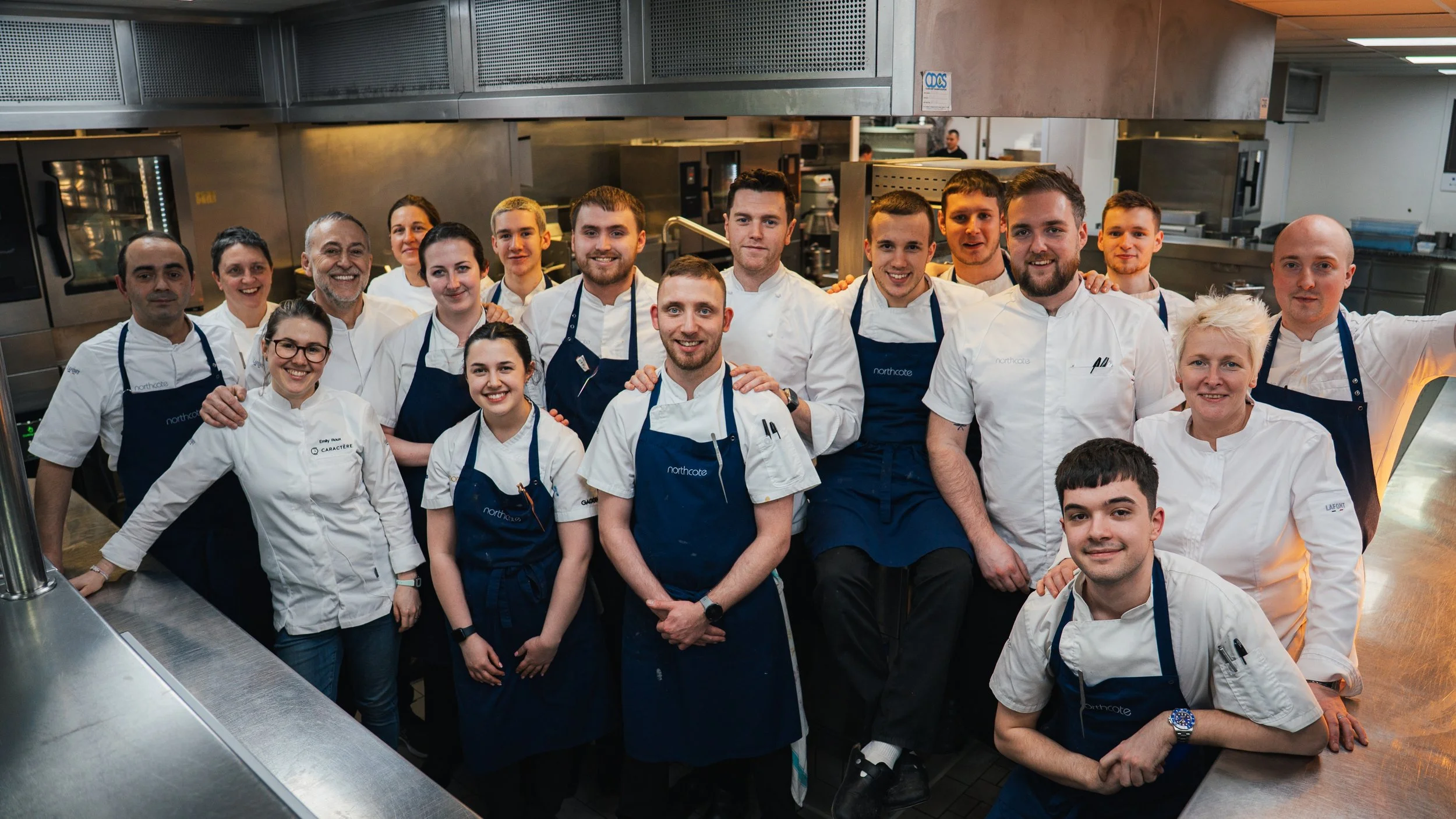 Group of chefs and kitchen staff in a professional kitchen, smiling for the camera.