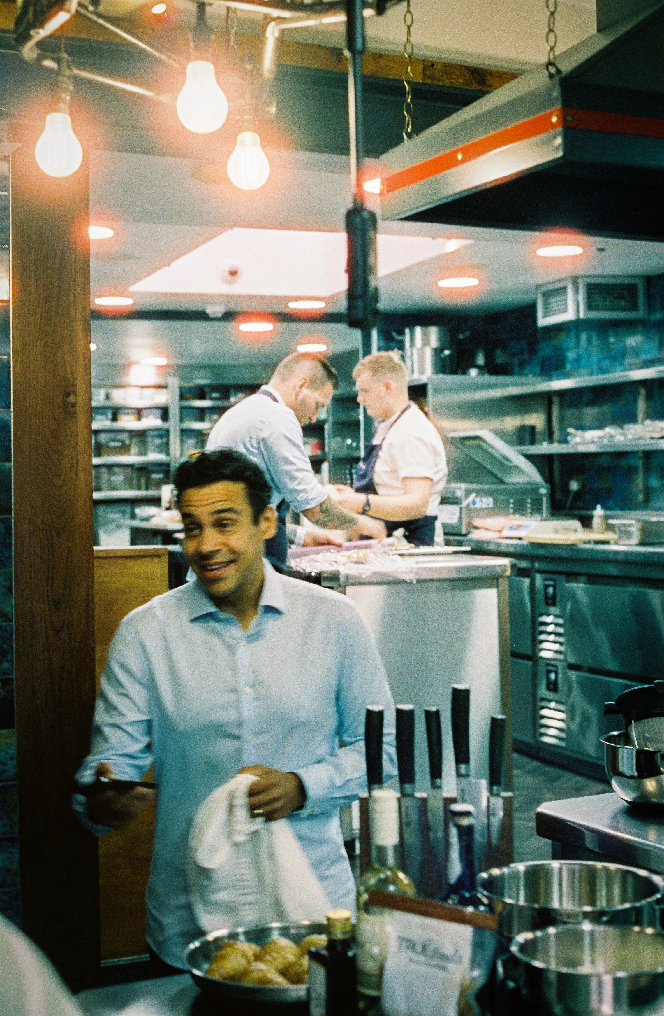 A scene in a restaurant kitchen showing two chefs preparing food, with a man in a blue shirt and a woman in a white shirt. The kitchen has stainless steel equipment, shelves, and cooking supplies. Warm lighting includes hanging light bulbs and recessed ceiling lights.