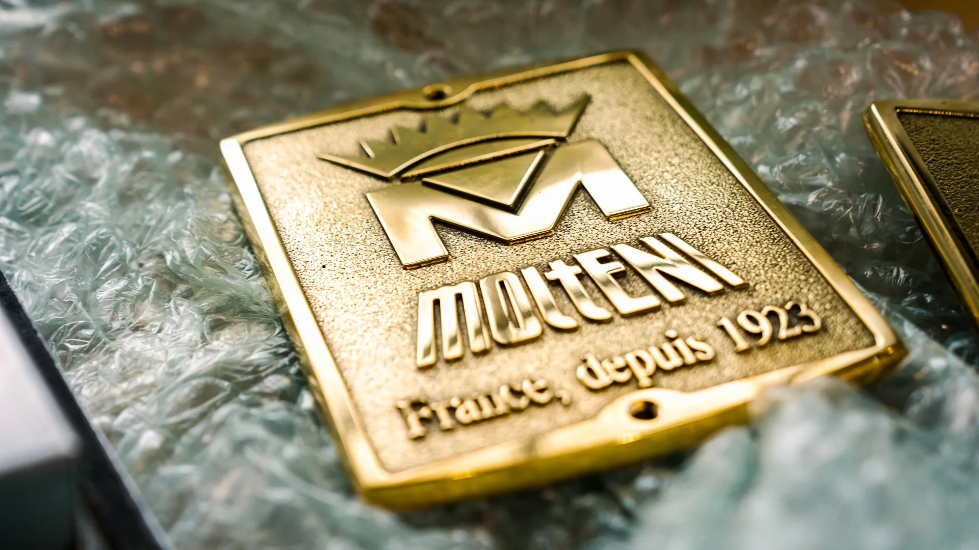 Close-up of a gold-colored badge with the logo and inscription for Maten, a French brand established in 1925, placed on a textured surface.