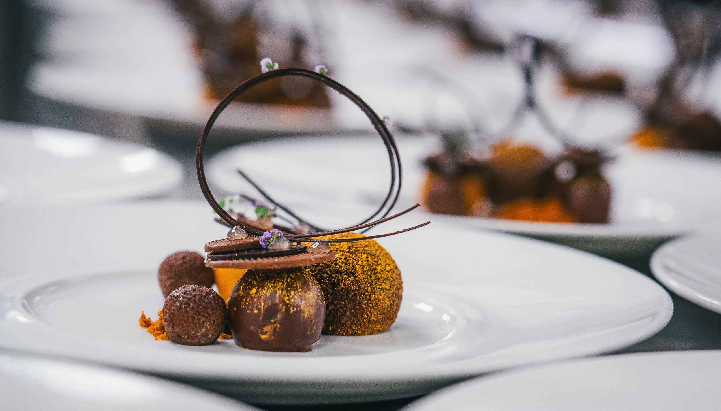 A plated gourmet dessert featuring chocolate spheres, small round truffles, and decorative edible elements topped with tiny purple flowers, garnished with vibrant orange dust, presented on a white plate.