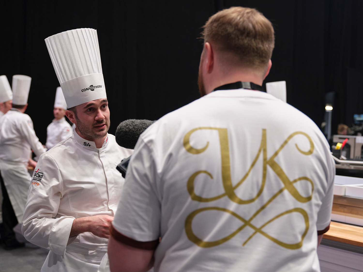 Two chefs in white uniforms and tall hats having a discussion at a kitchen event. Several other chefs are in the background, and one person is filming with a microphone.