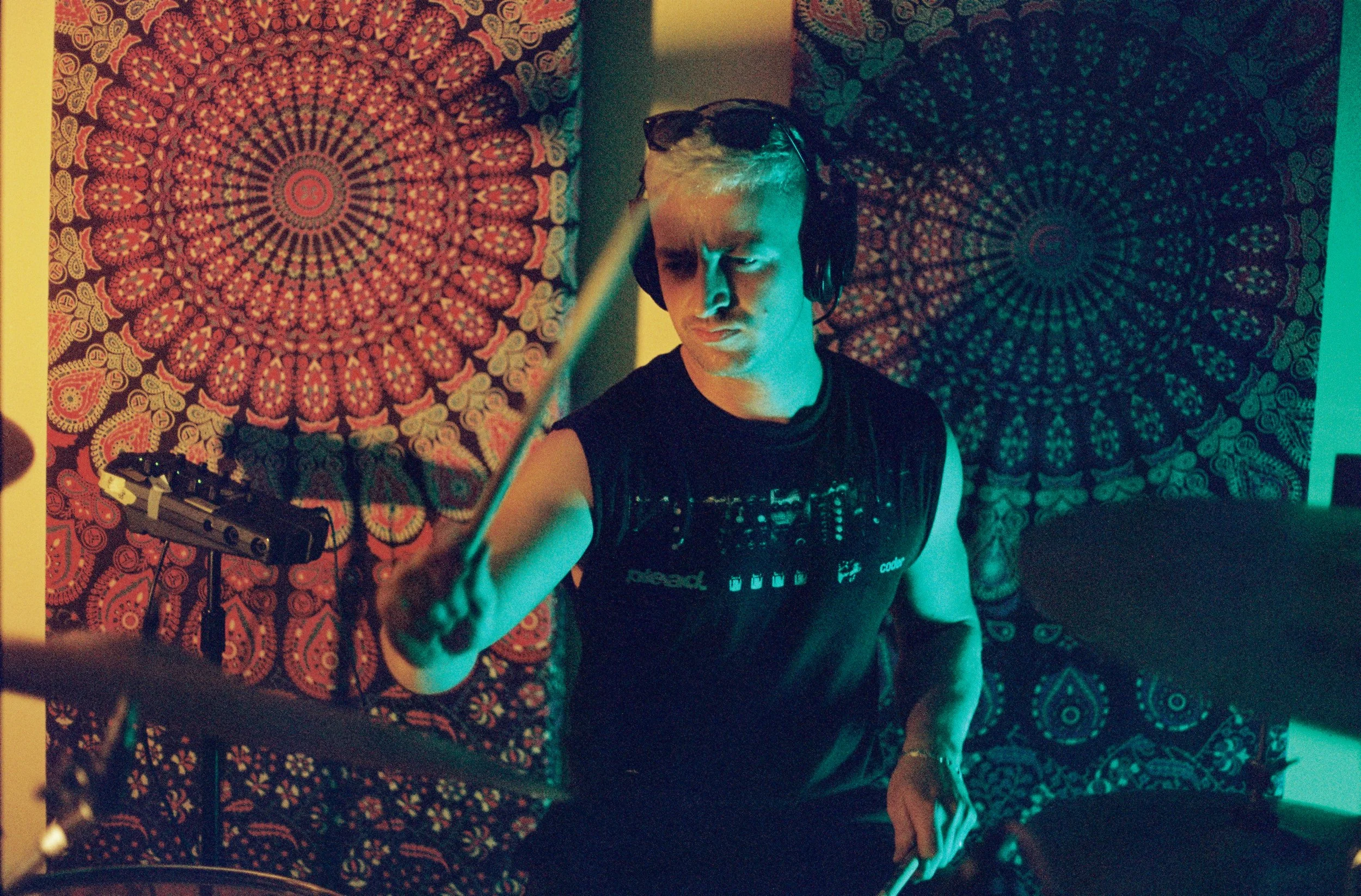 A DJ wearing black sunglasses and a black sleeveless shirt with music icons, is adjusting sound equipment in a room decorated with colorful, intricate mandala tapestry. The room is lit with teal and dim yellow lighting.