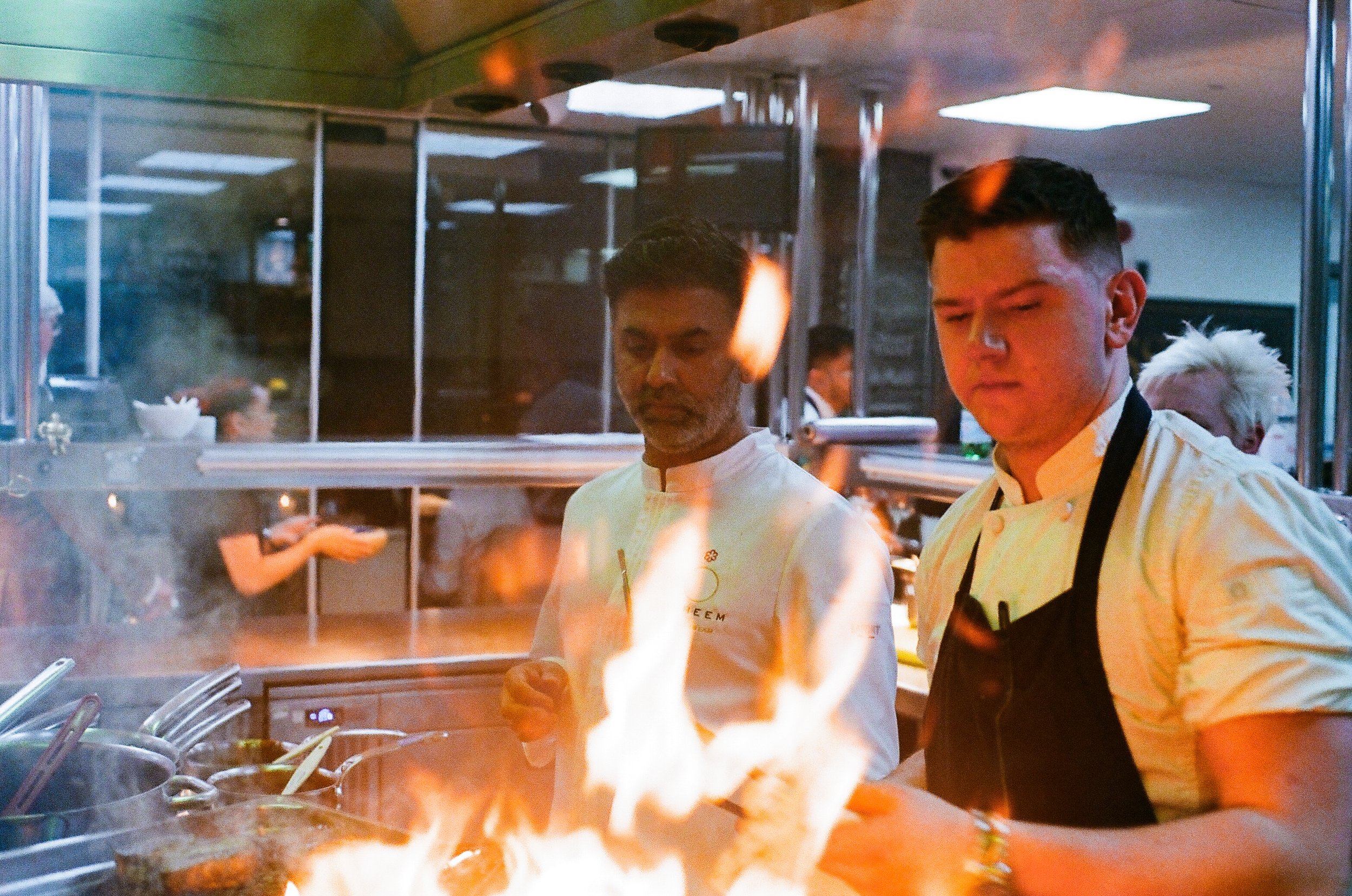 Two chefs working in a busy kitchen, focused on a flaming dish.