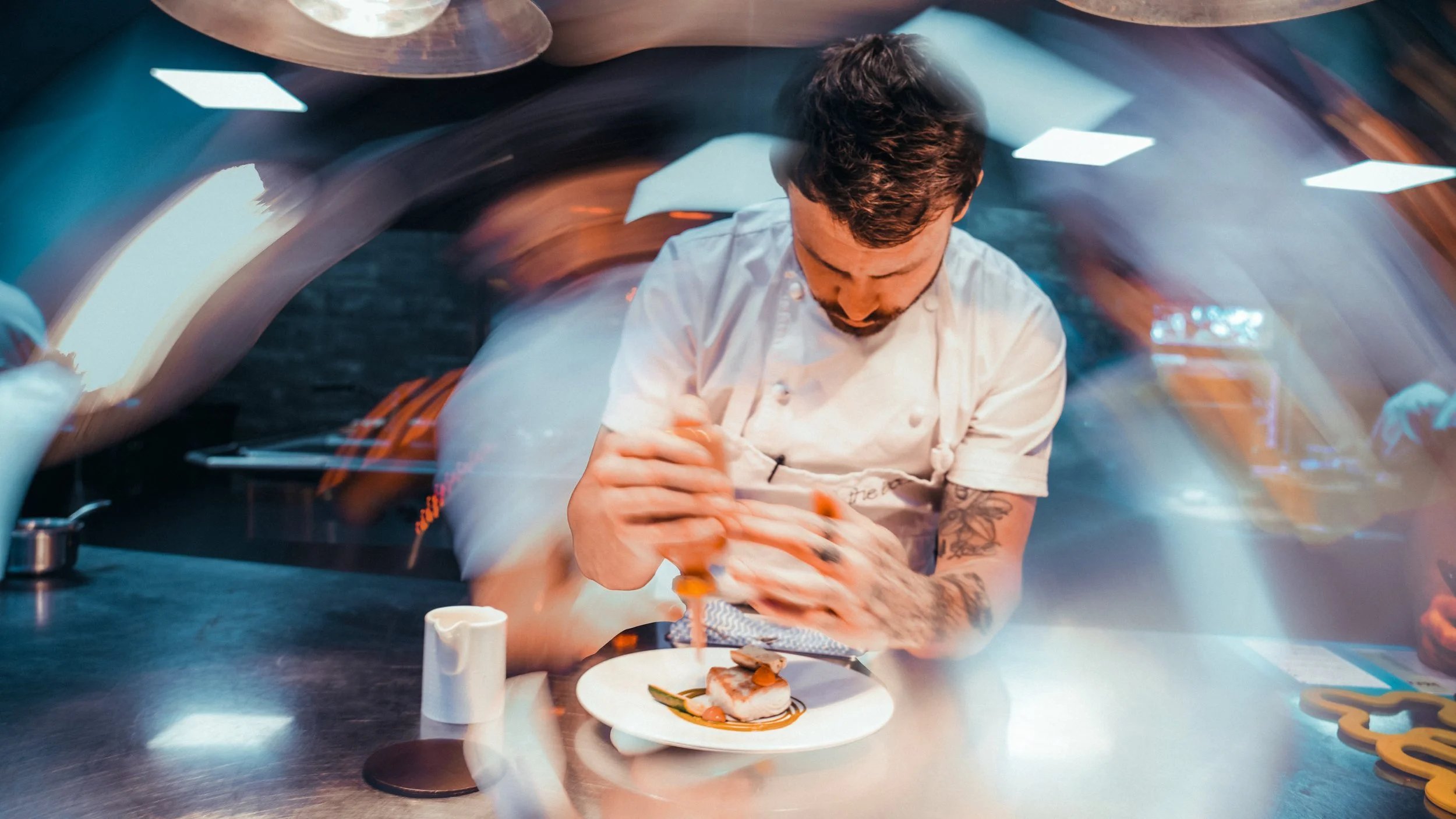 A chef in a white uniform with tattoos on his arms is preparing food in a modern kitchen with colorful lighting and contemporary decor.