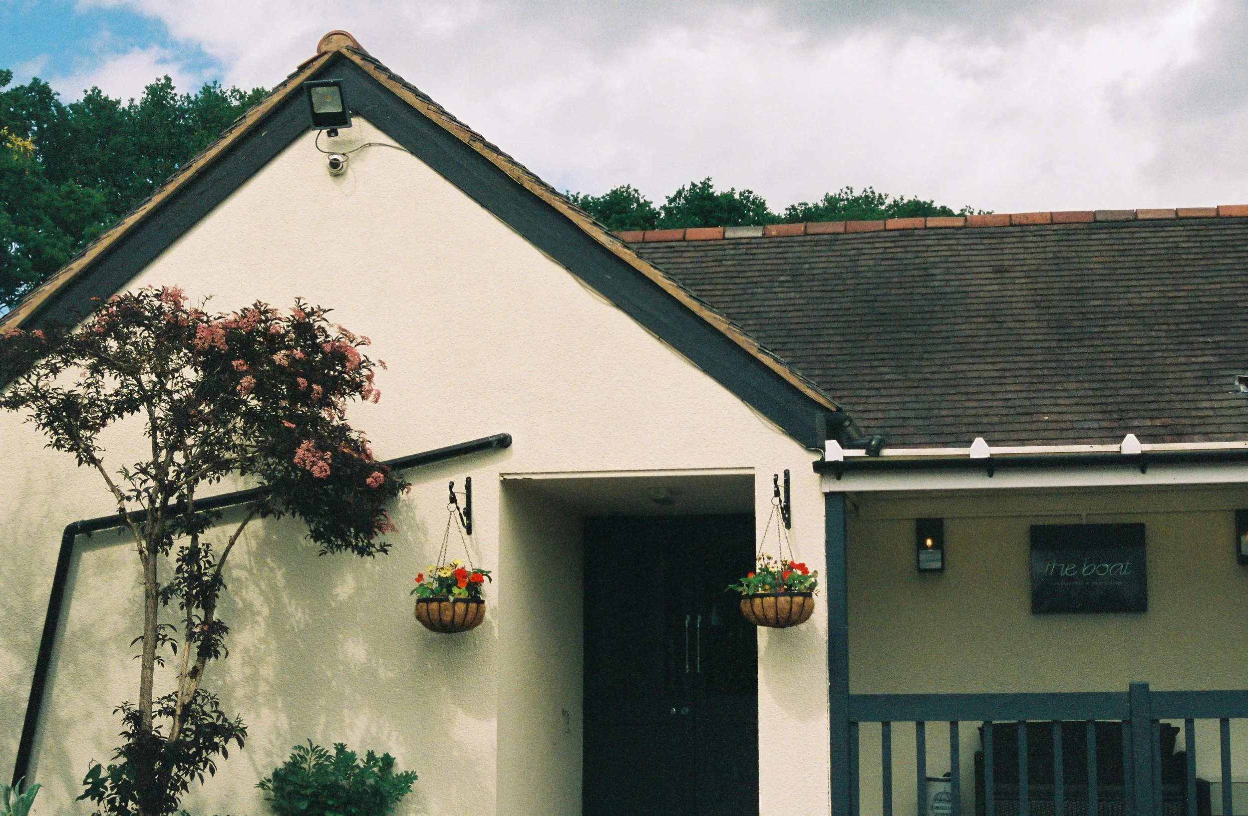 A house with a steep gabled roof, white exterior walls, and hanging flower baskets on the porch. There is a small tree with pink flowers and greenery in front of the house. The sky is overcast with clouds.