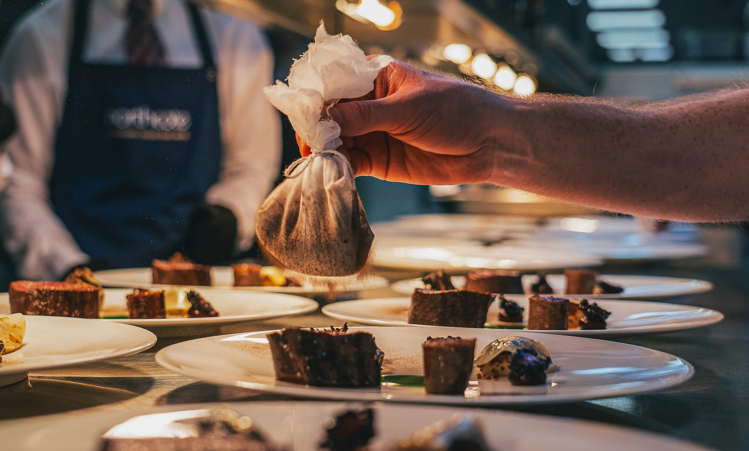A person is placing a dessert on a white plate in a professional kitchen setting. Multiple plates with similar desserts are lined up on a stainless steel counter.