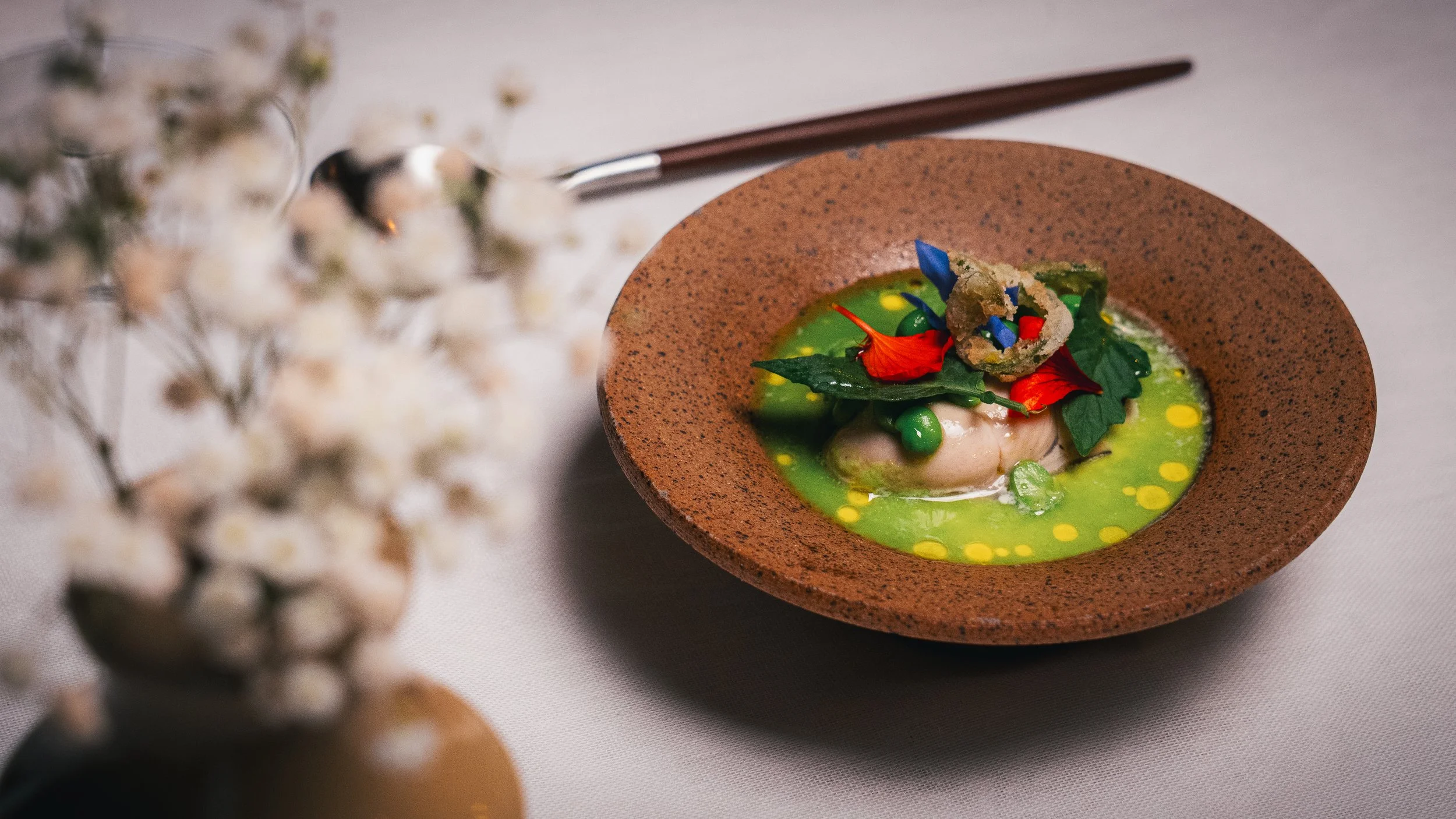 A gourmet dish featuring a poached or lightly cooked protein, green peas, leafy greens, and edible flowers in a green sauce, served in a rustic brown bowl on a white tablecloth, with a spoon and chopsticks nearby, partially blurred white flowers in f