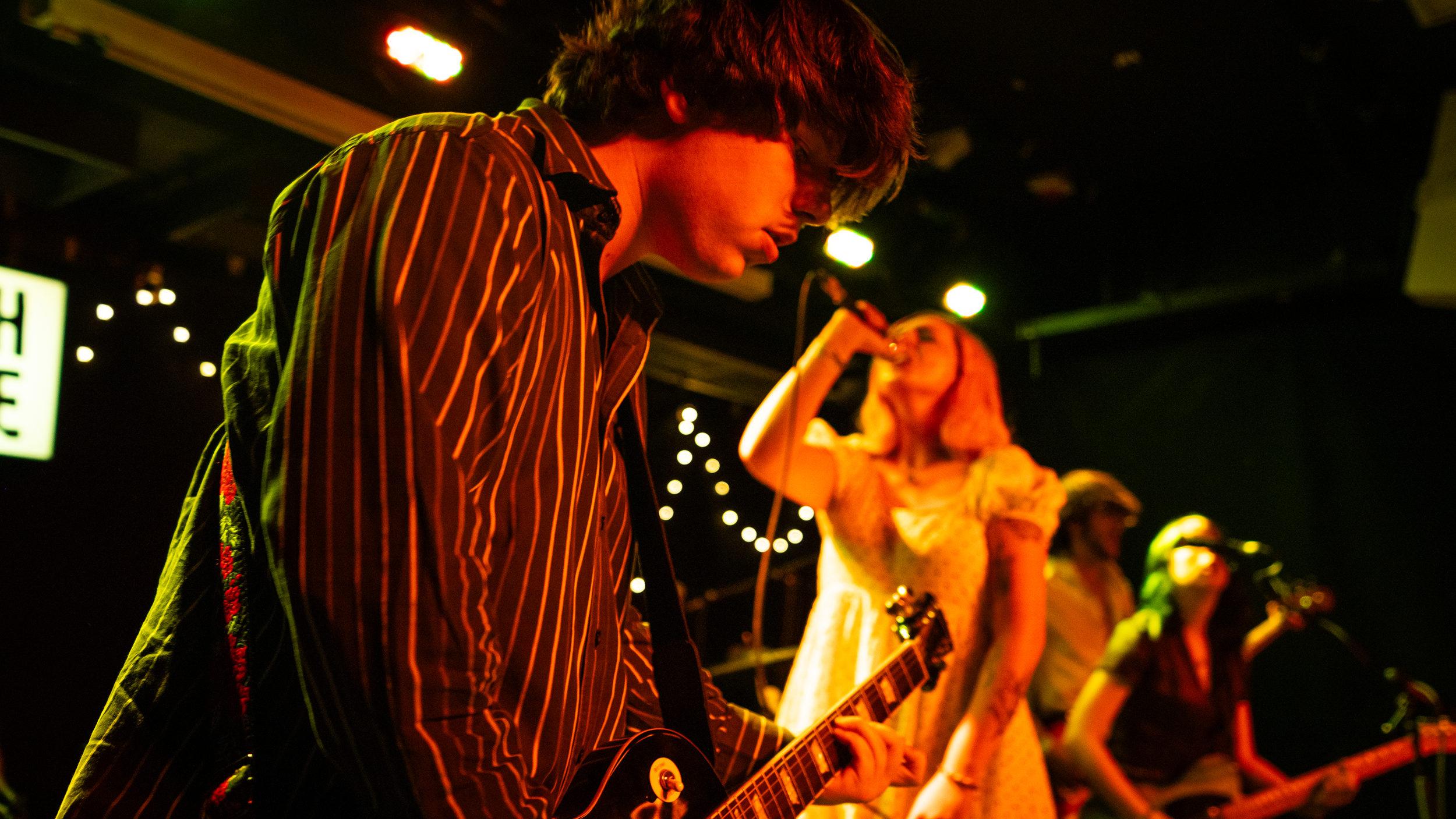 Musicians performing on stage with a guitarist in the foreground and a vocalist in the background under stage lights.