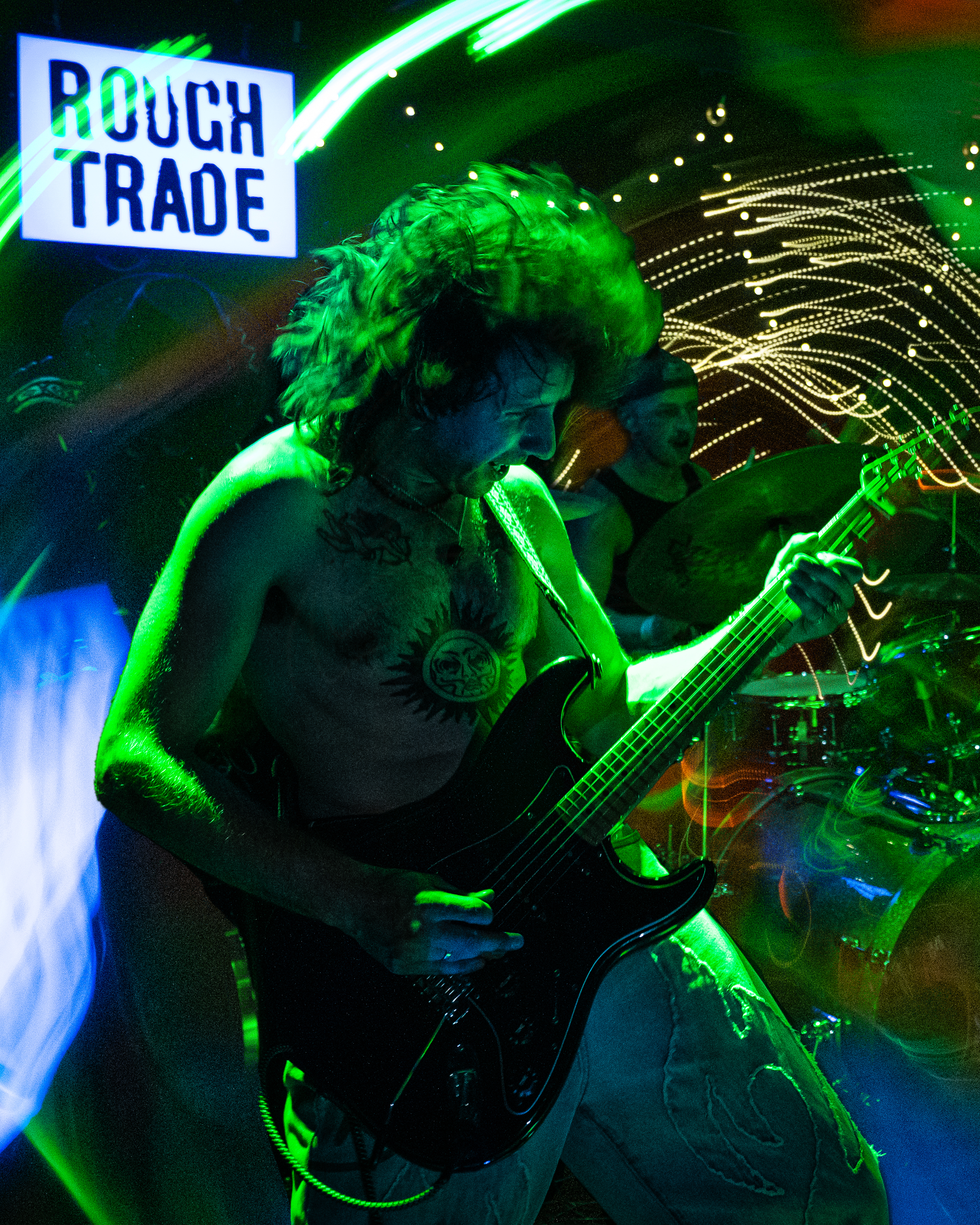 A woman with tattoos playing an electric guitar in a dimly lit room illuminated by green neon and colorful light streaks, with a sign in the background that reads 'Rough Trade'.