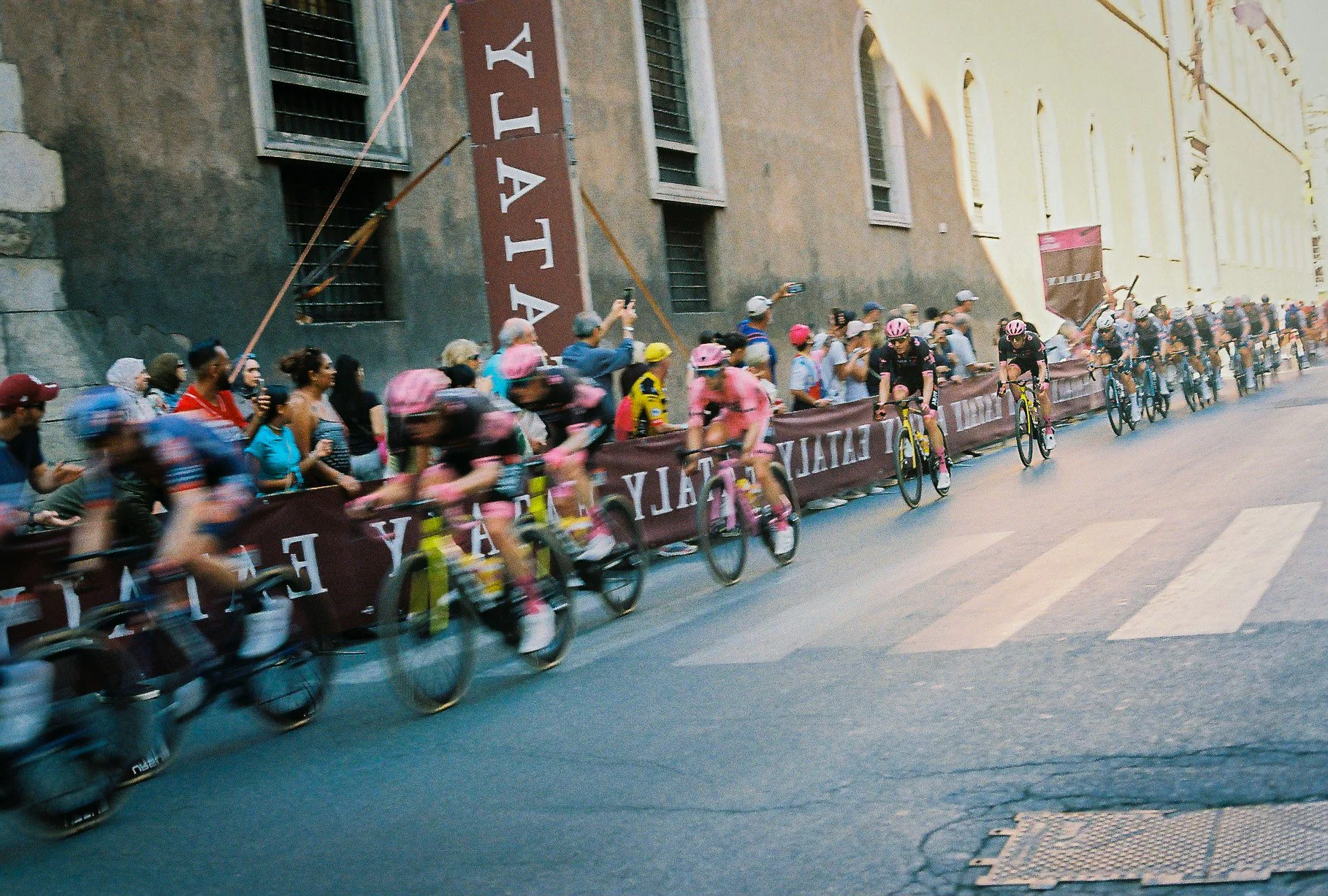 A group of cyclists racing on a city street, with spectators watching from the sidewalk and taking photos.