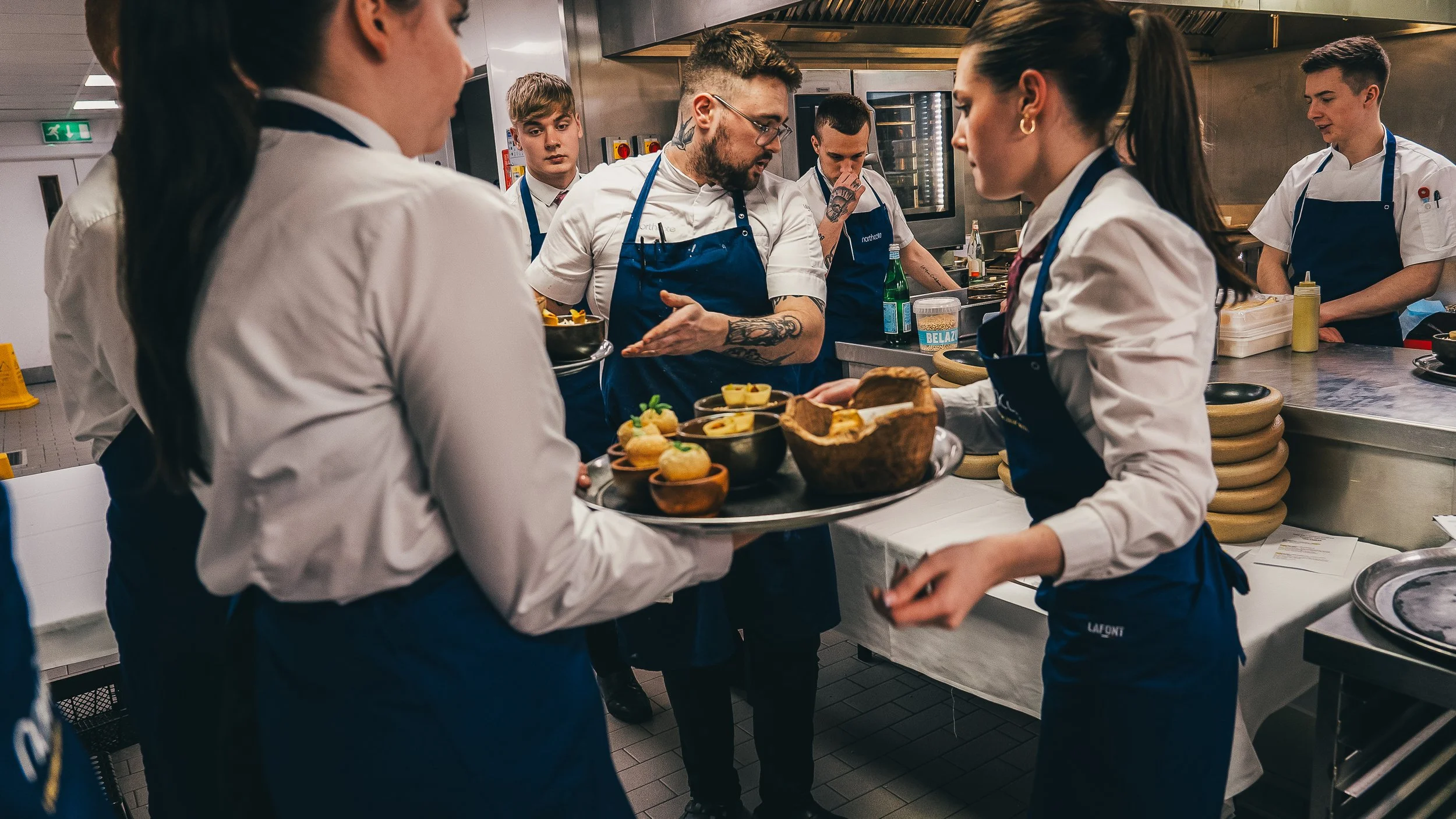 Group of chefs and kitchen staff in a professional kitchen preparing and serving food, with plating, utensils, bottles, and ingredients visible.