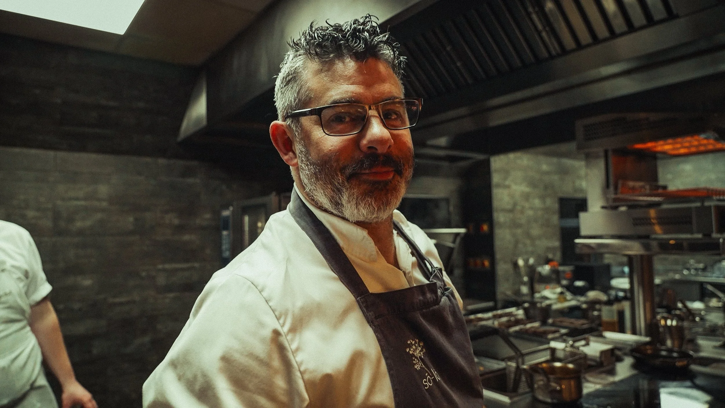 A man with glasses and a beard wearing a white chef's jacket and black apron in a professional kitchen.