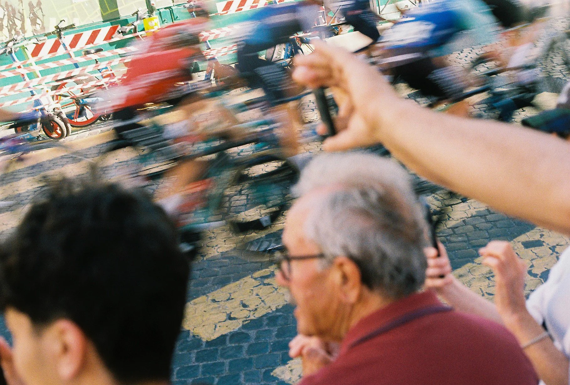 Blurred image of people riding bicycles in a city street, with a person in the foreground holding a dog and another person wearing glasses and a maroon shirt.